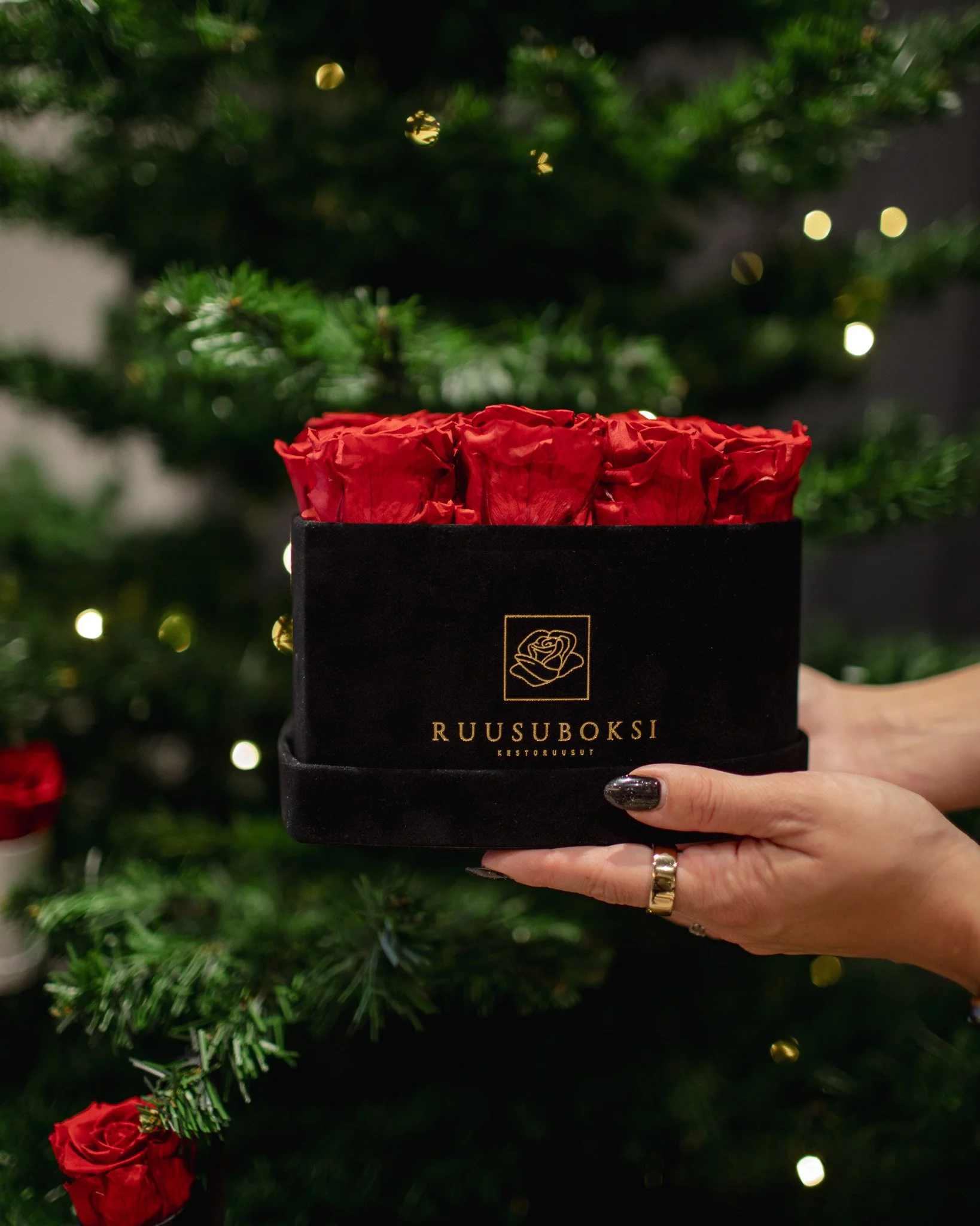 Käsin pidettävä musta lahjapakkaus, jossa on punaisia ruusuja ja teksti "RUUSUBOKSI KESTORUUSUT".