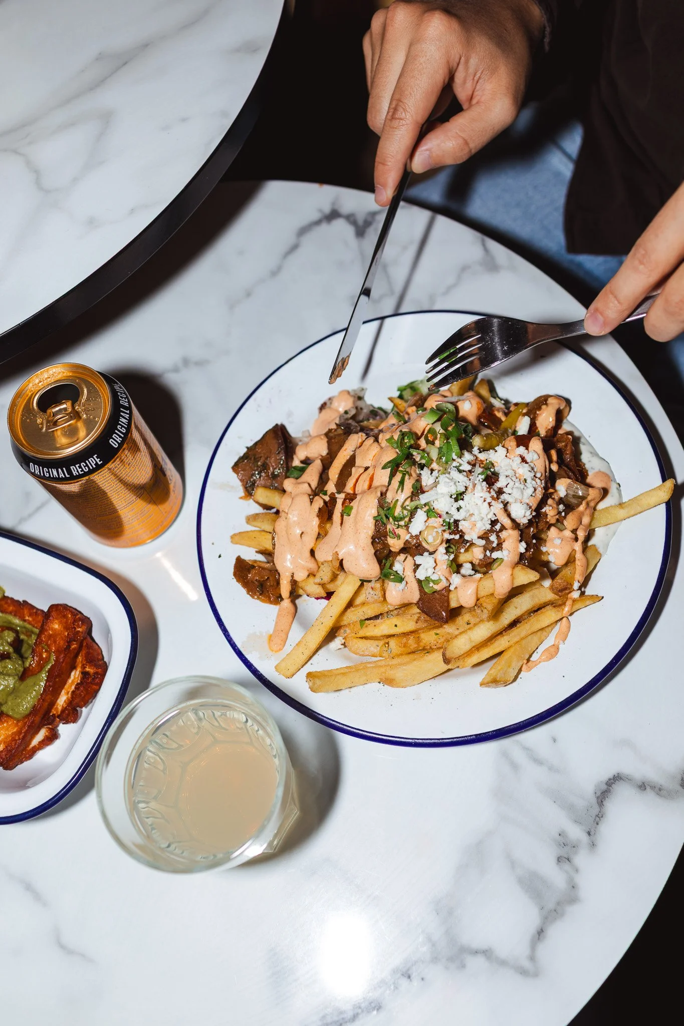 Henkilö syö ranskalaisia perunoita kastikkeella ja raastetulla juustolla pöydässä, jossa on myös juomaa ja muita ruokia.