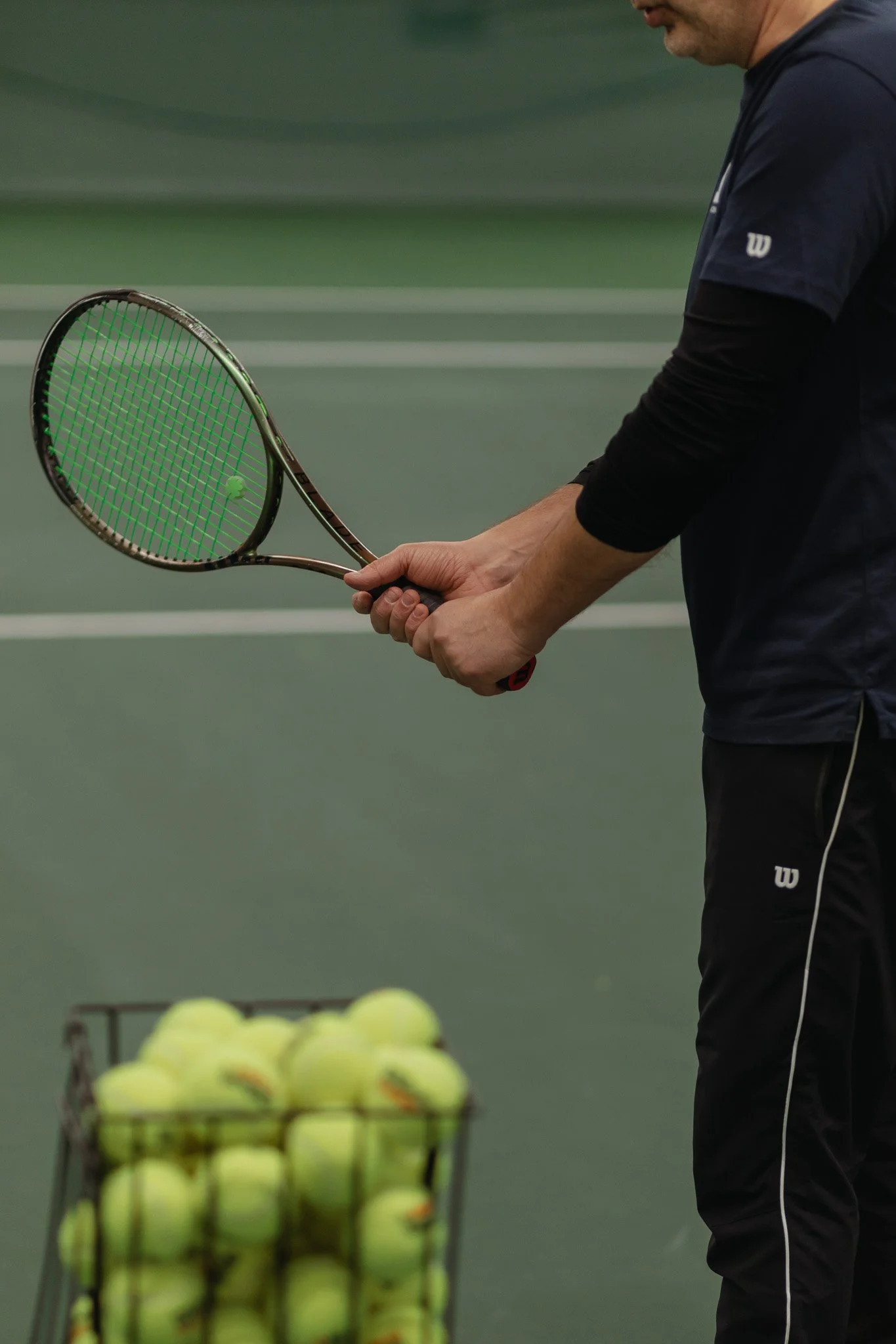 Mies kädessä tennismaila harjoittelee tennistä tenniskentällä, taustalla on kori täynnä uusia tennispalloja.