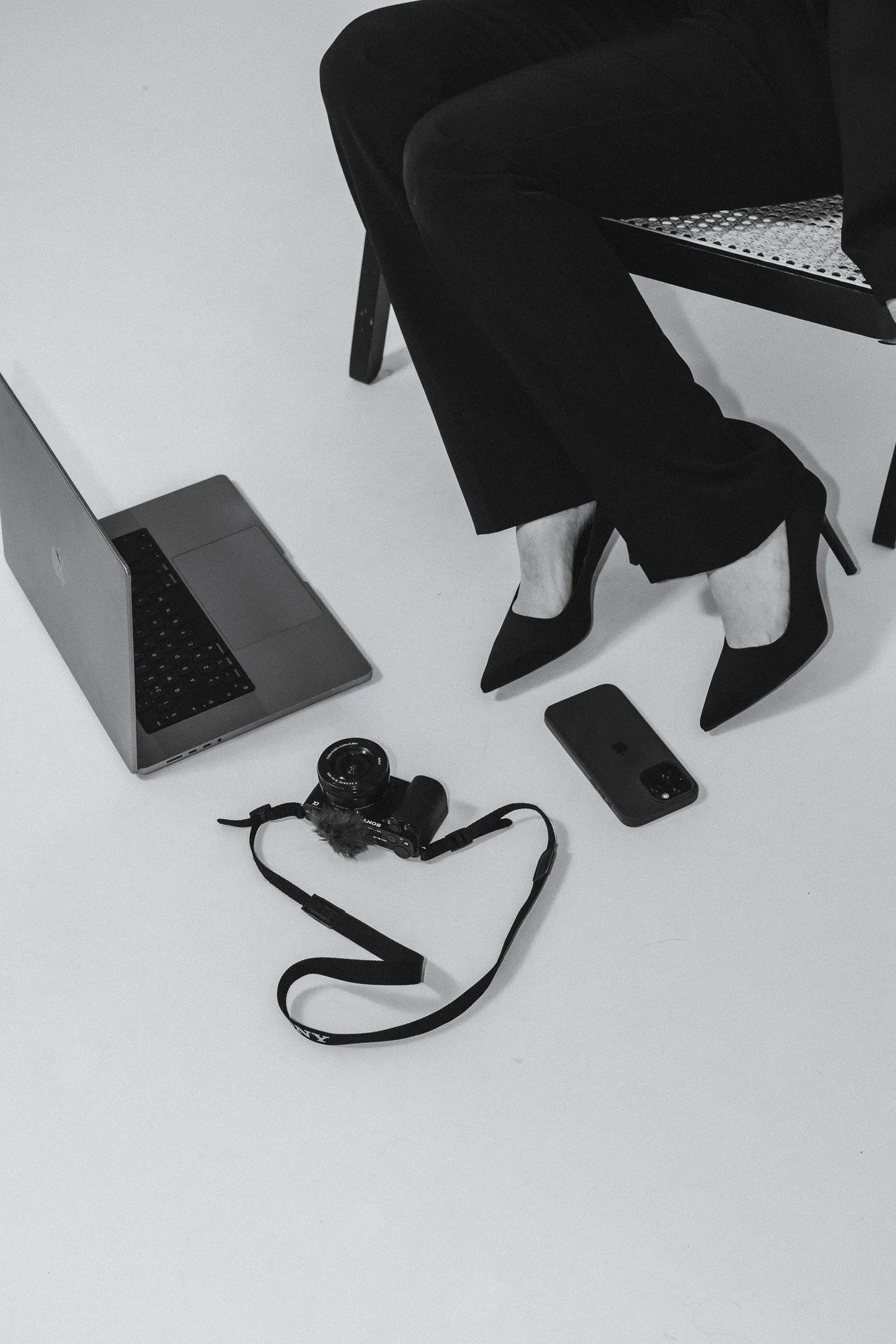 Mustavalkoinen kuva henkilöstä, joka istuu tuolilla, hänellä on kiinalainen tyyli ja korolliset kengät, lähellä on kannettava tietokone, kamera ja älypuhelin.