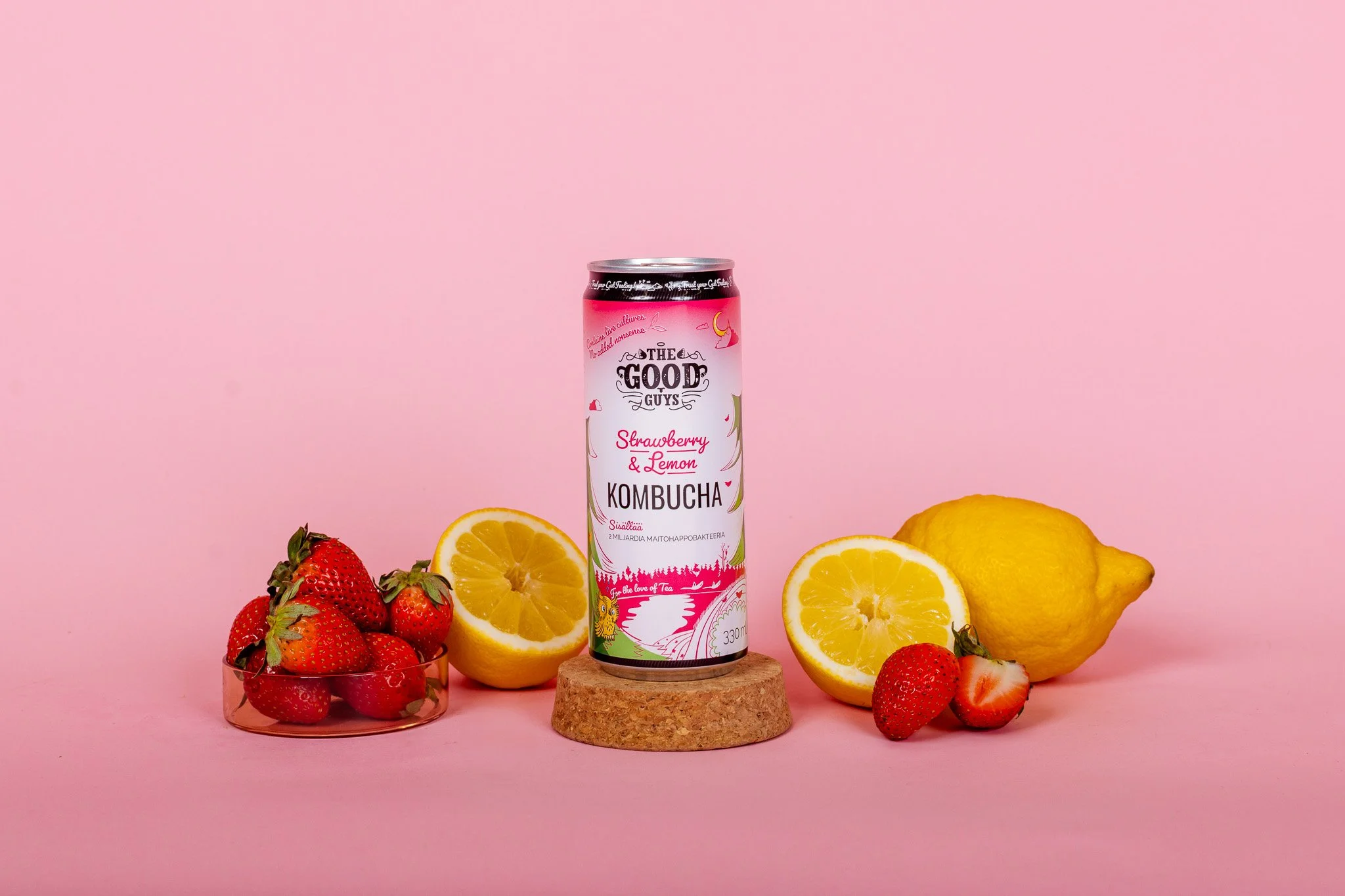Kuvassa on tölkki The Good Guys Strawberry & Lemon Kombucha keskellä, ympärillä tuoreita mansikoita, puolikurkkua ja sitruunoita, osa sitruunoista on puolitetuina, ja tölkki on asetettu pienen puupalan päälle vaaleanpunaisella taustalla.