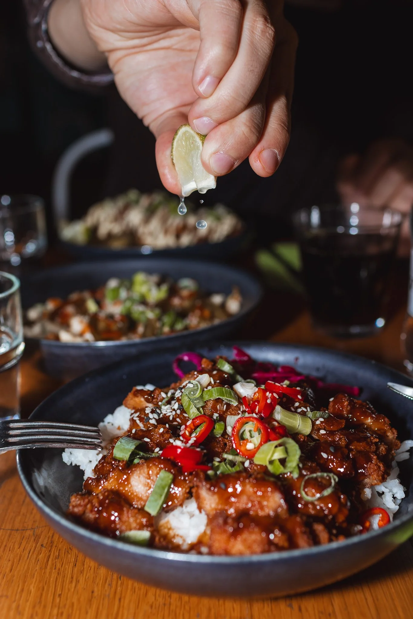 Lähikuva aasialaisesta ruoasta lautasella, jossa on riisiä, paistettua kanaa ja vihanneksia, ja henkilö puristaa limonimouskaa ruokaan.