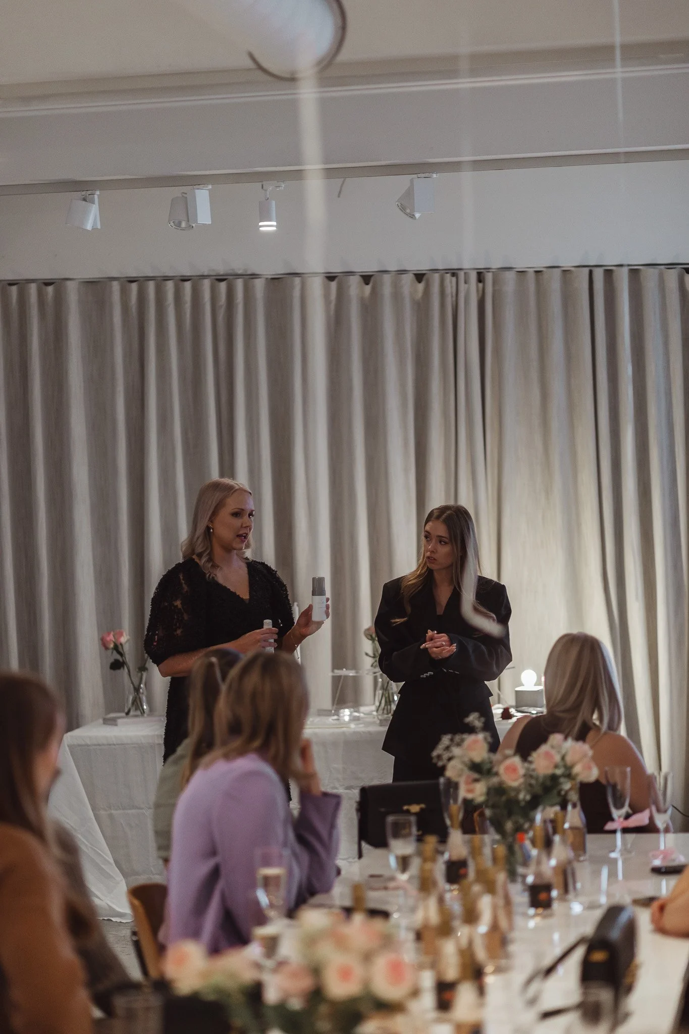 Nainen esittää tuotteita ystävälleen juhlapöydän ääressä, juhlaväkeä pöydän äärellä.