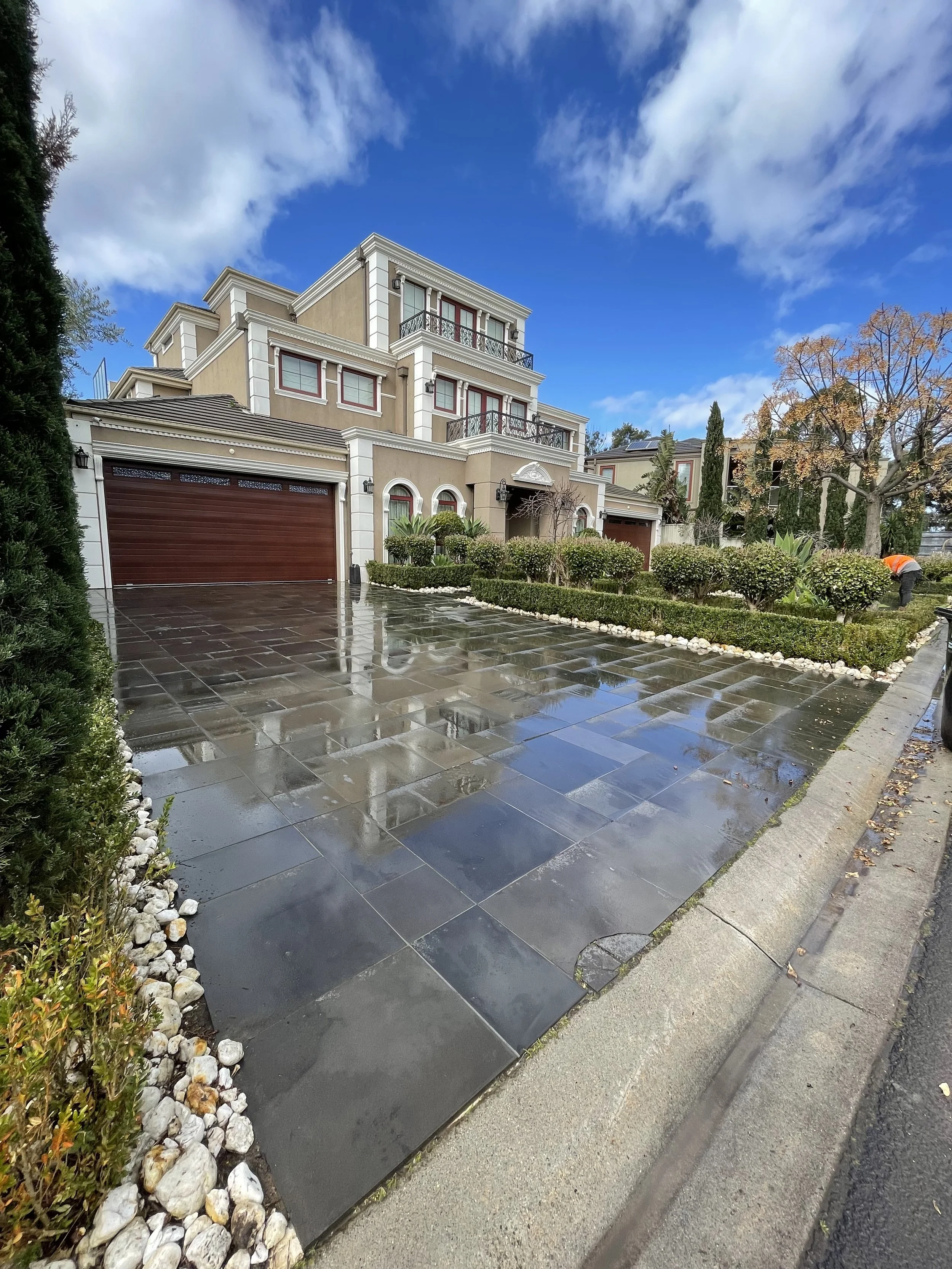 A wet driveway in front of a multi-story house with beige walls, white trim, and red accents under a mostly cloudy sky.