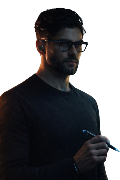 A man with dark hair, glasses, and a beard writes with a pen on a surface, wearing a dark shirt, against a plain dark background.