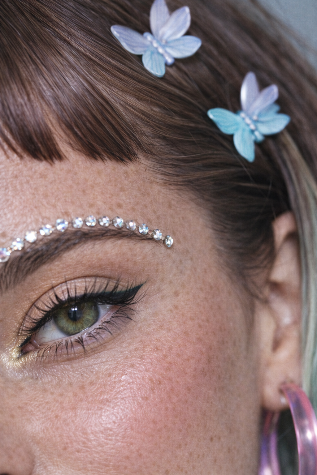 Close-up of a woman's eye with makeup, decorated with small crystals above the eyebrow, and two blue butterfly-shaped hair clips in red-brown hair with platinum streaks. Custom AI model, AI influencer.