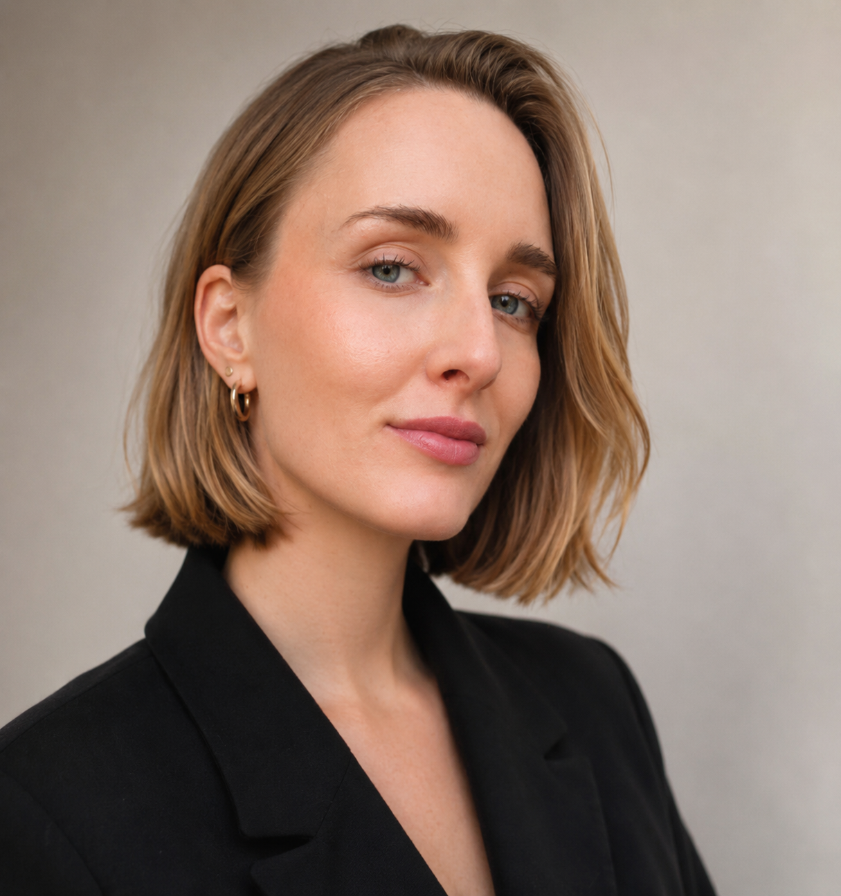 A woman with short, wavy, light brown hair, blue eyes, wearing a black blazer and gold hoop earrings, looking confidently at the camera against a plain, neutral background.