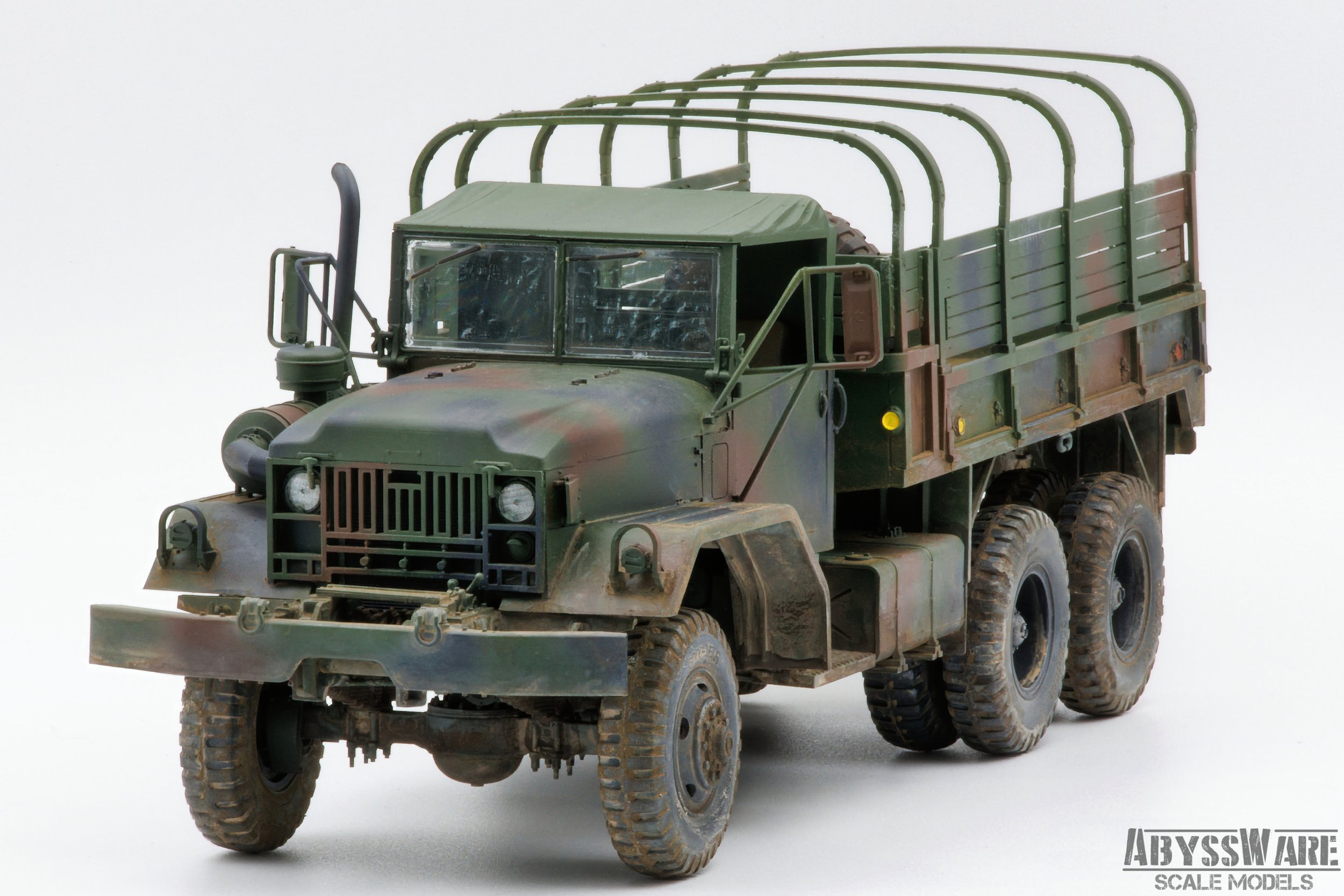 Scale model of a military cargo truck with a camouflage paint job, featuring six large tires, a cab with a windshield, and a cargo bed with a metal railing, placed on a plain white background.
