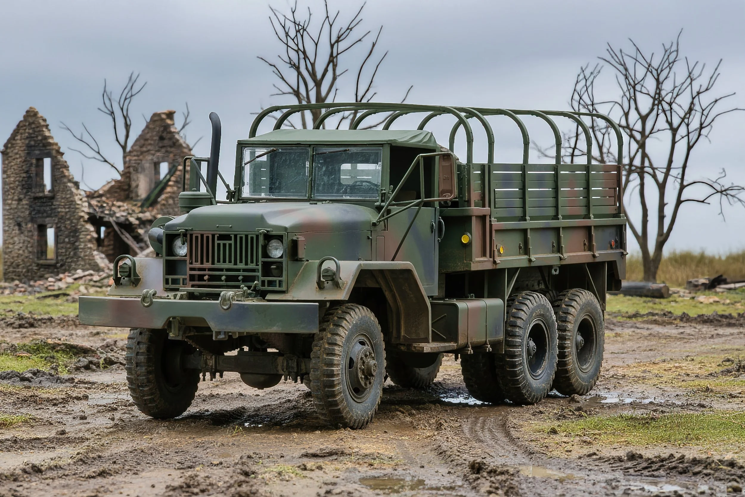 A military truck with camouflage paint parked on muddy ground, with ruined building and leafless trees in the background.