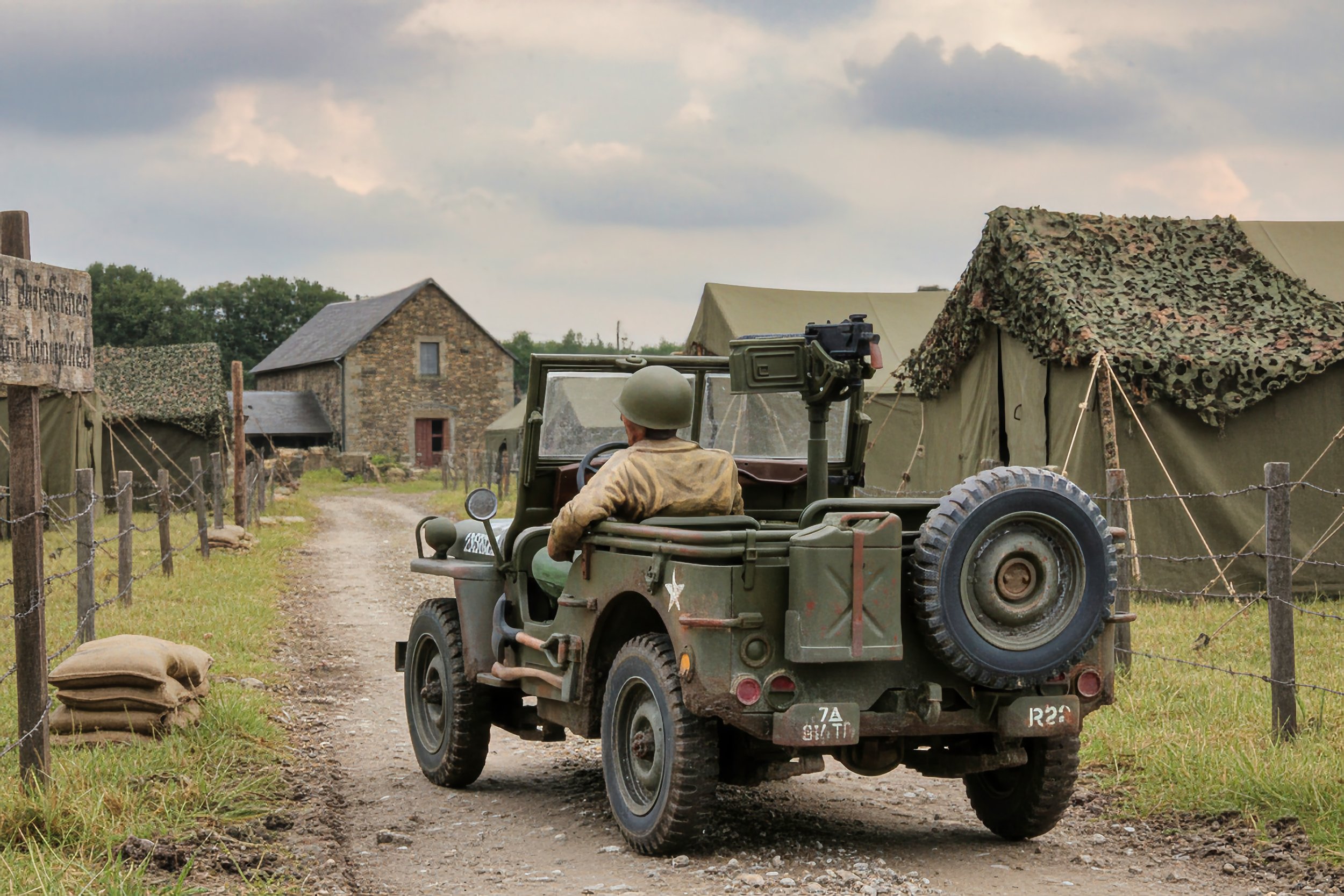 A soldier driving a vintage military jeep on a dirt road in a rural, wartime camp with military tents and a stone building under a cloudy sky.