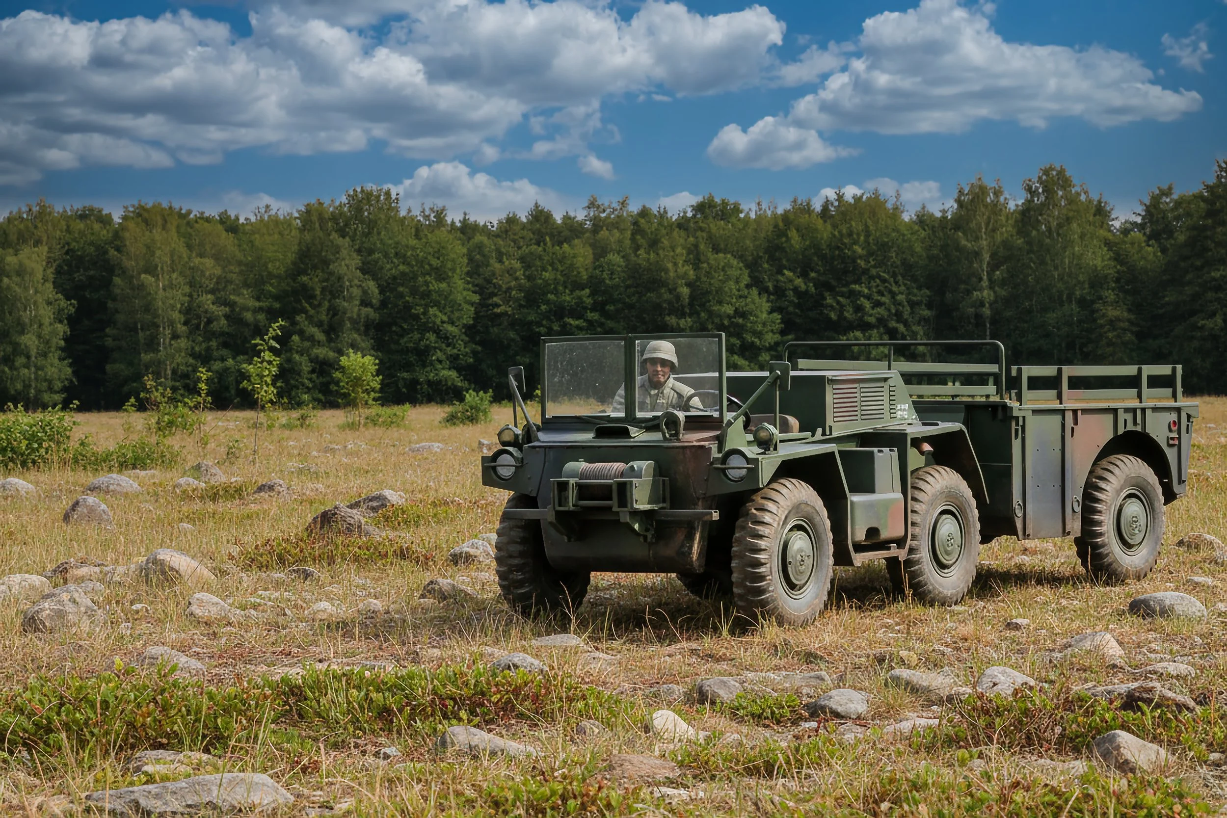 A military-style all-terrain vehicle with a person wearing a hat inside, parked in a grassy field with rocks and trees in the background under a partly cloudy sky.