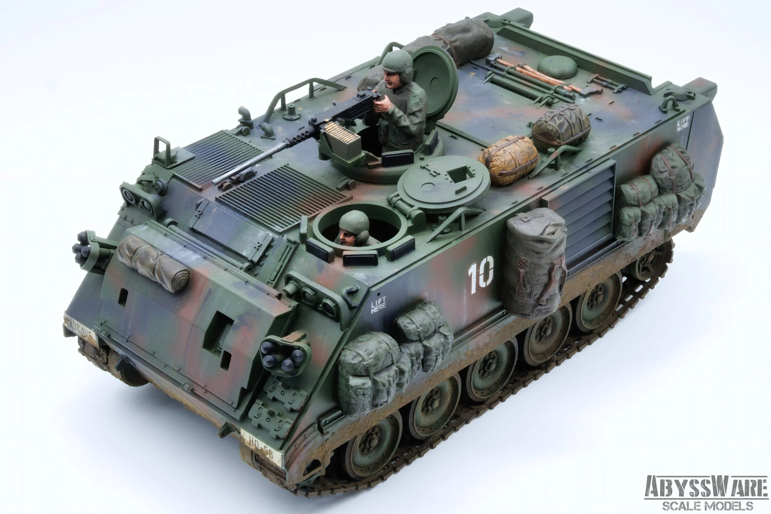 A detailed scale model of a military armored vehicle with camouflage paint, featuring two soldiers figurines on top, one manning a machine gun and a woman peering out from a hatch. The vehicle is equipped with various bags and gear attached on the si