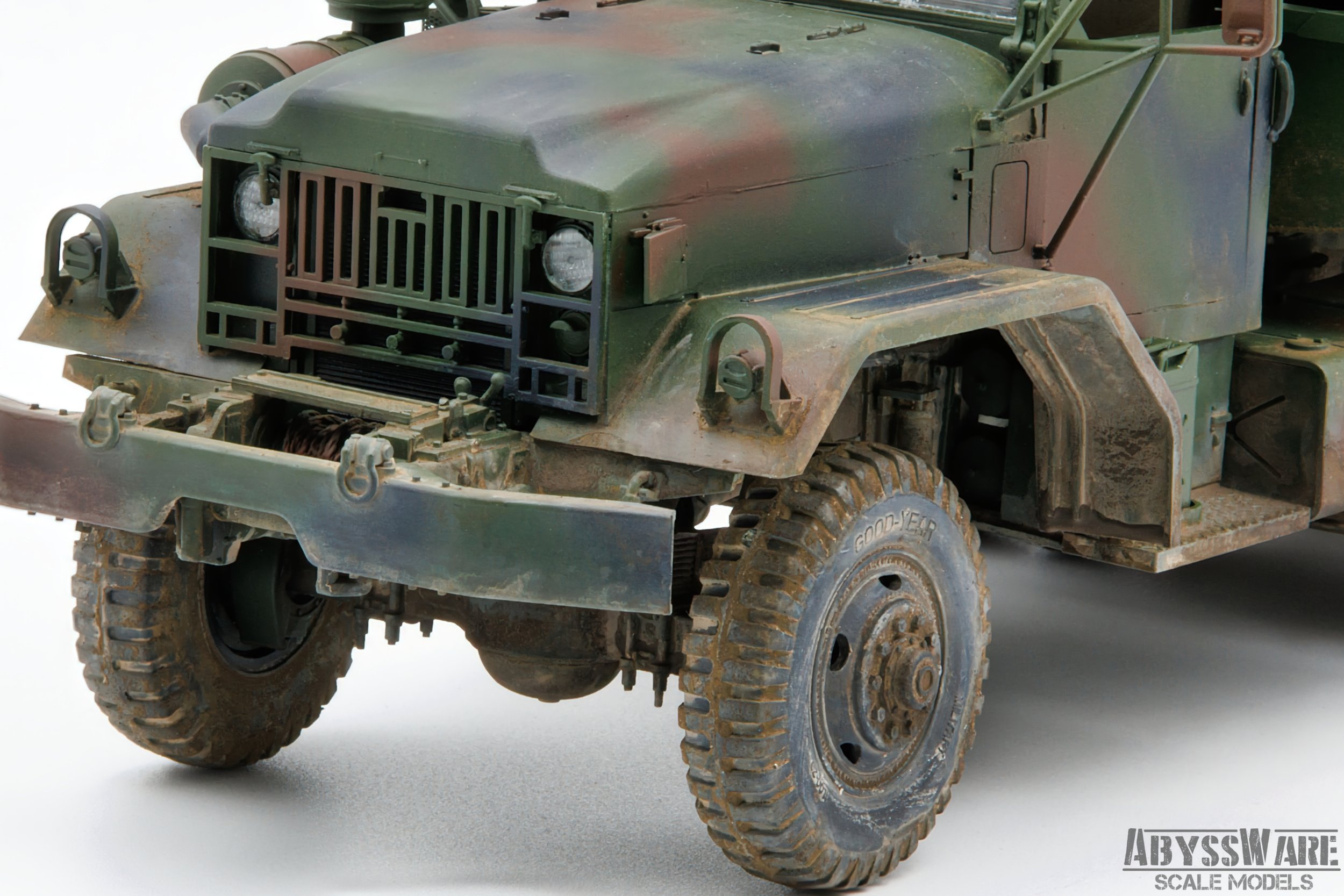 Close-up of a weathered military truck with a green camouflage paint job and large tires, showing detailed front grille and bumper, on a plain white background.