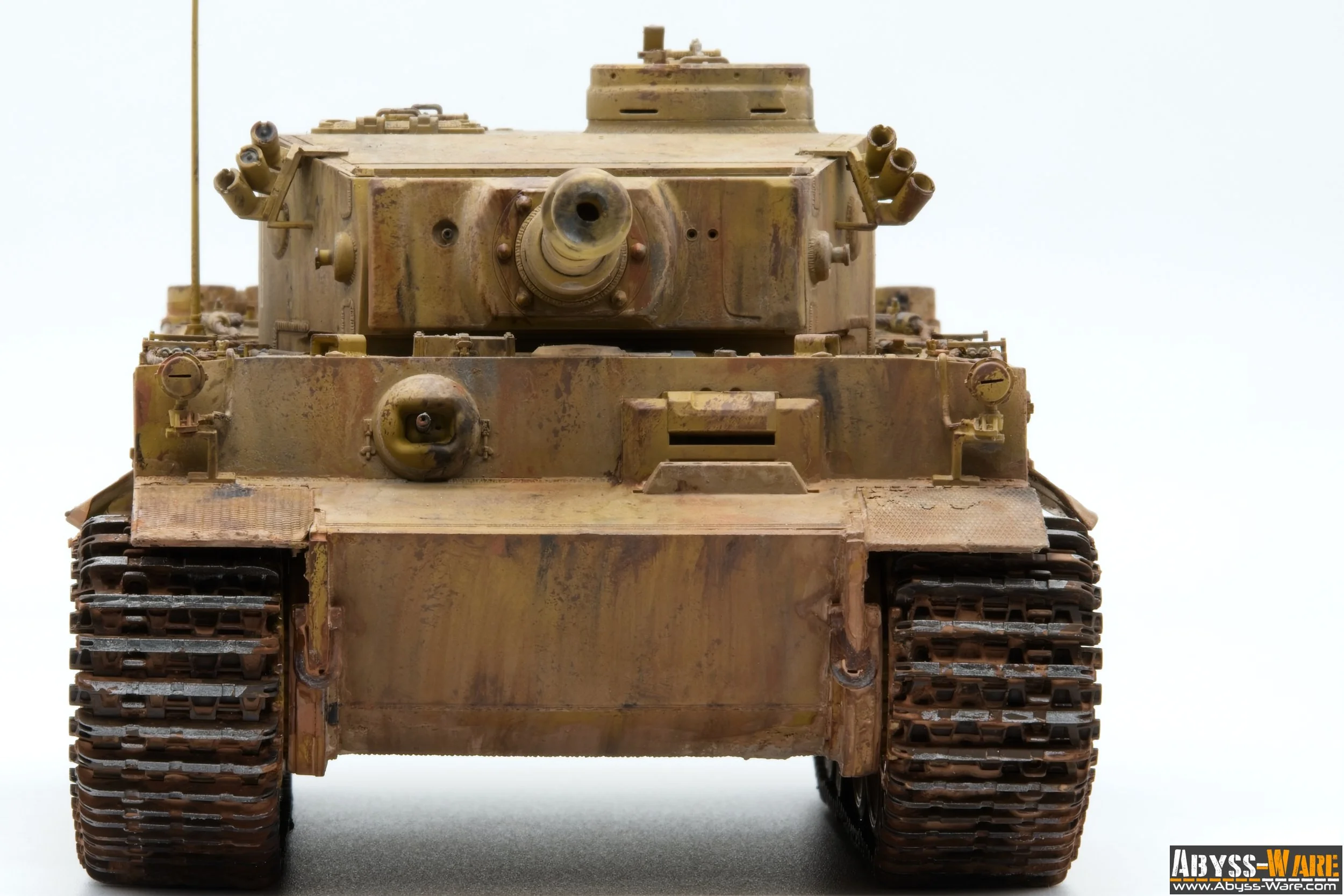 A front view of a weathered, desert-colored military tank with a large main gun barrel pointing forward, set against a plain, light background.