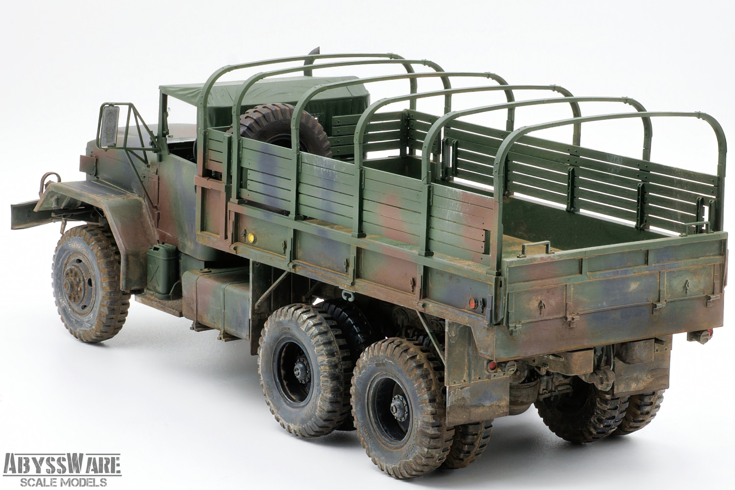 Scale model of a military cargo truck with a camouflage paint job, six wheels, open flatbed with side rails, and a spare tire mounted on the bed.