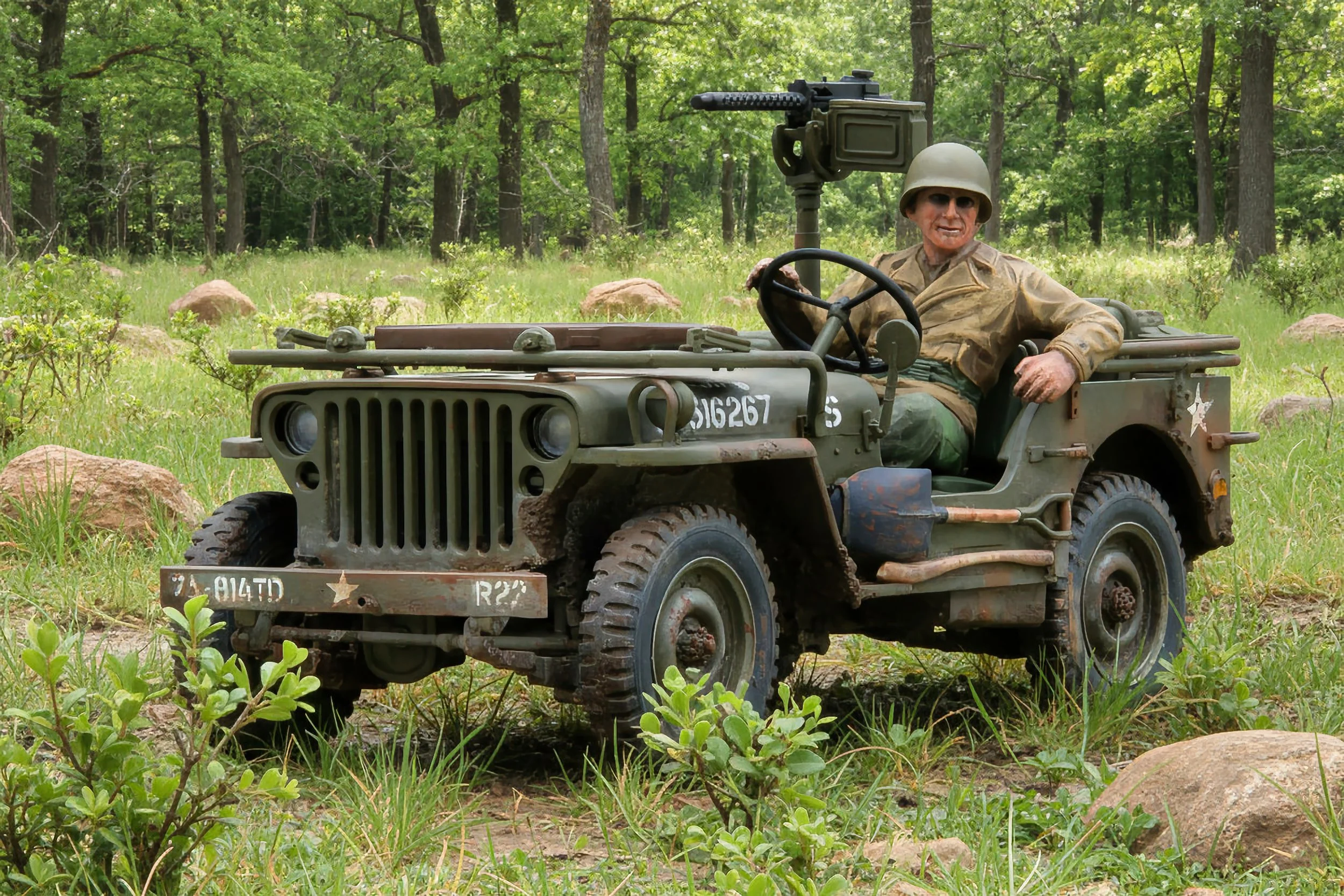 A person dressed in military gear sitting in a vintage military jeep with a mounted machine gun, in a wooded outdoor setting.