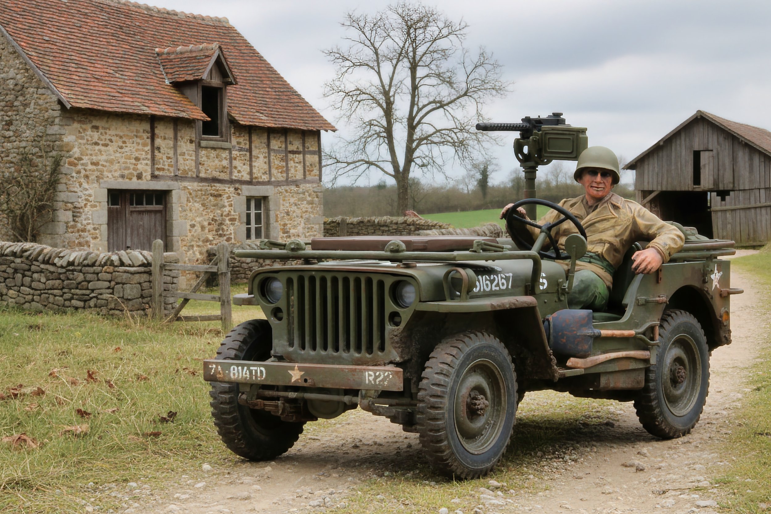 A person dressed in military uniform sitting in a vintage military jeep with a mounted gun, in front of a stone and wood farmhouse under a cloudy sky.
