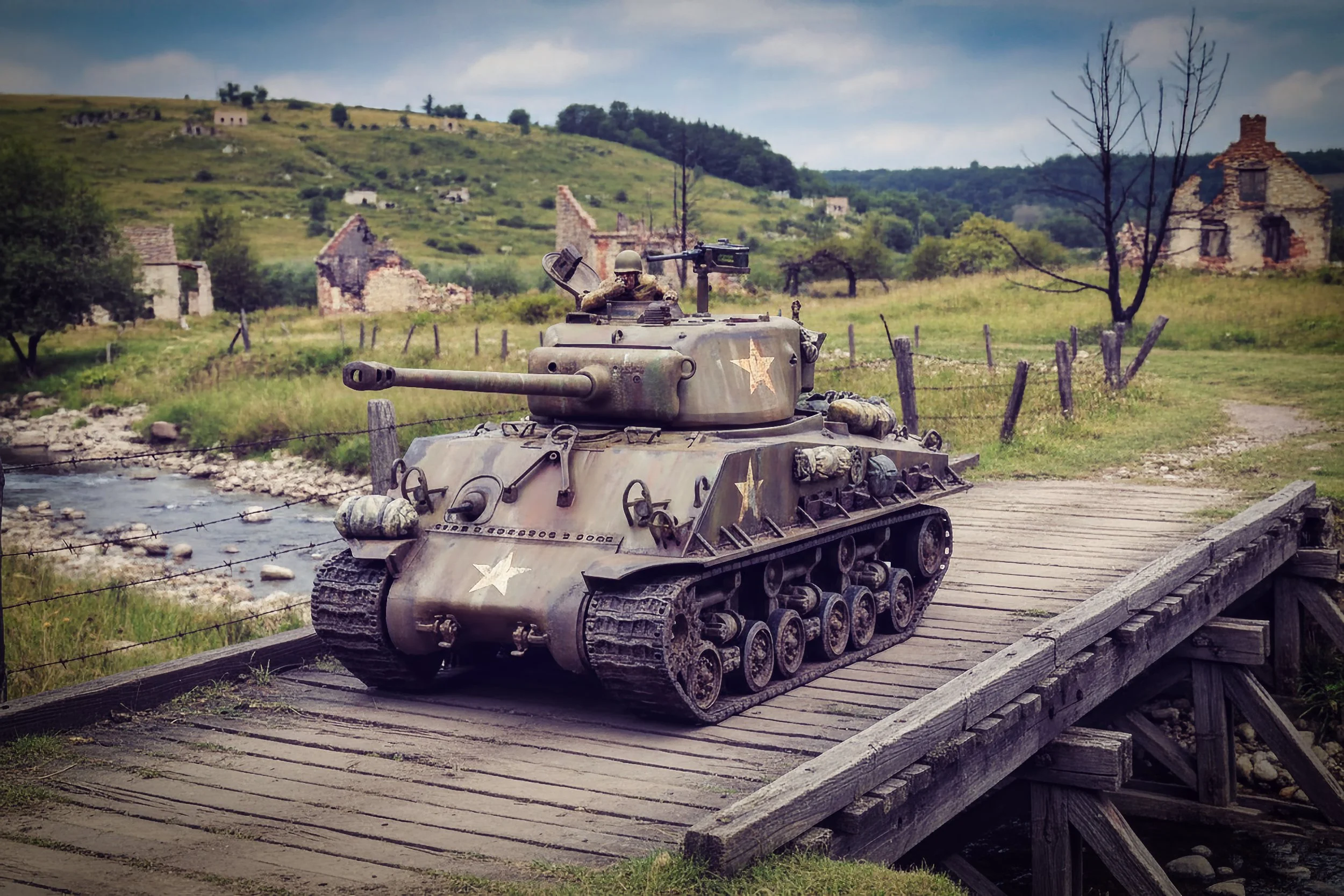 A military tank on a wooden bridge in a rural landscape with hills, old buildings, and a creek in the background.