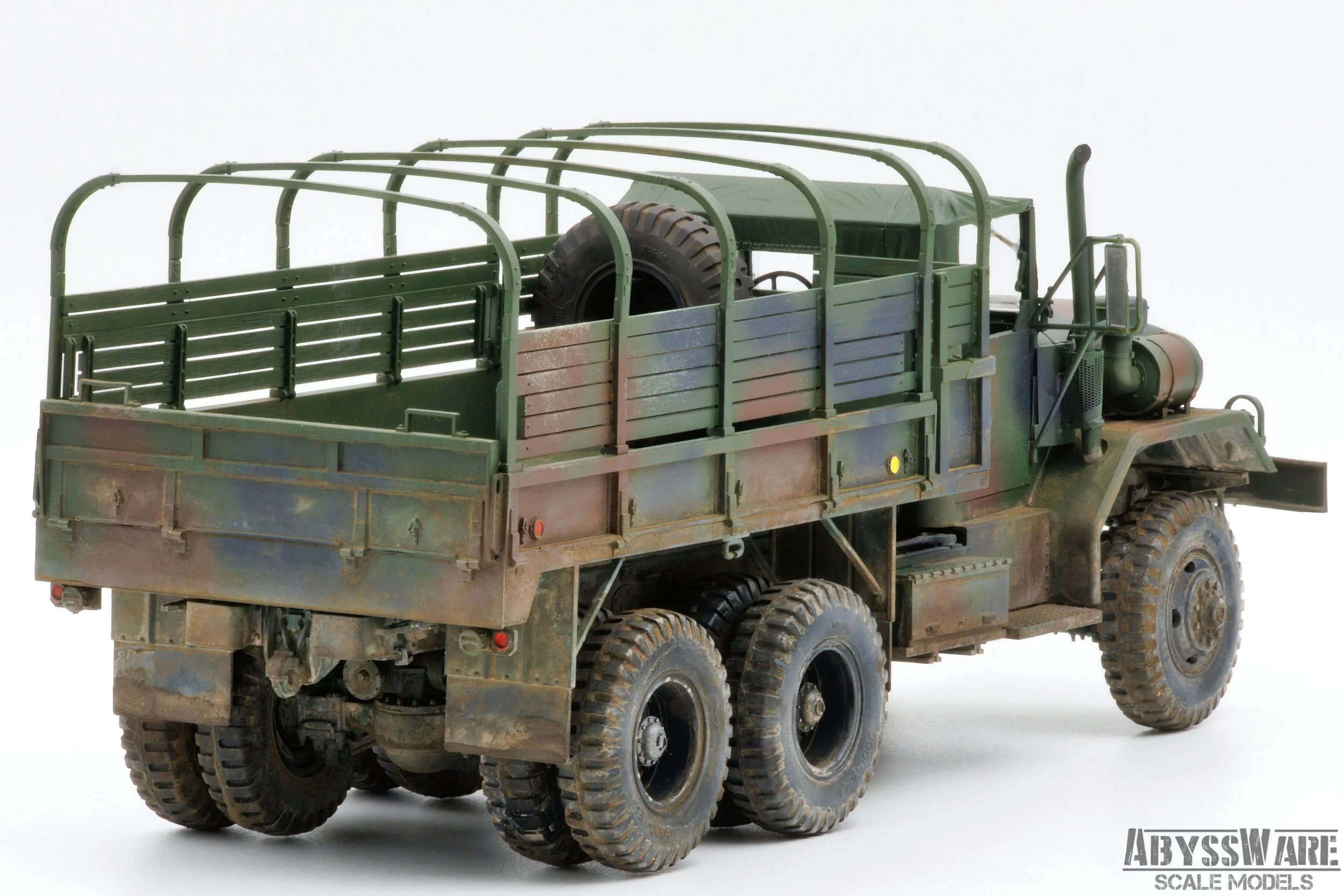 A miniature model of a military truck with six wheels, painted in a green camouflage pattern, with a spare tire mounted on the bed and metal railings along the sides.