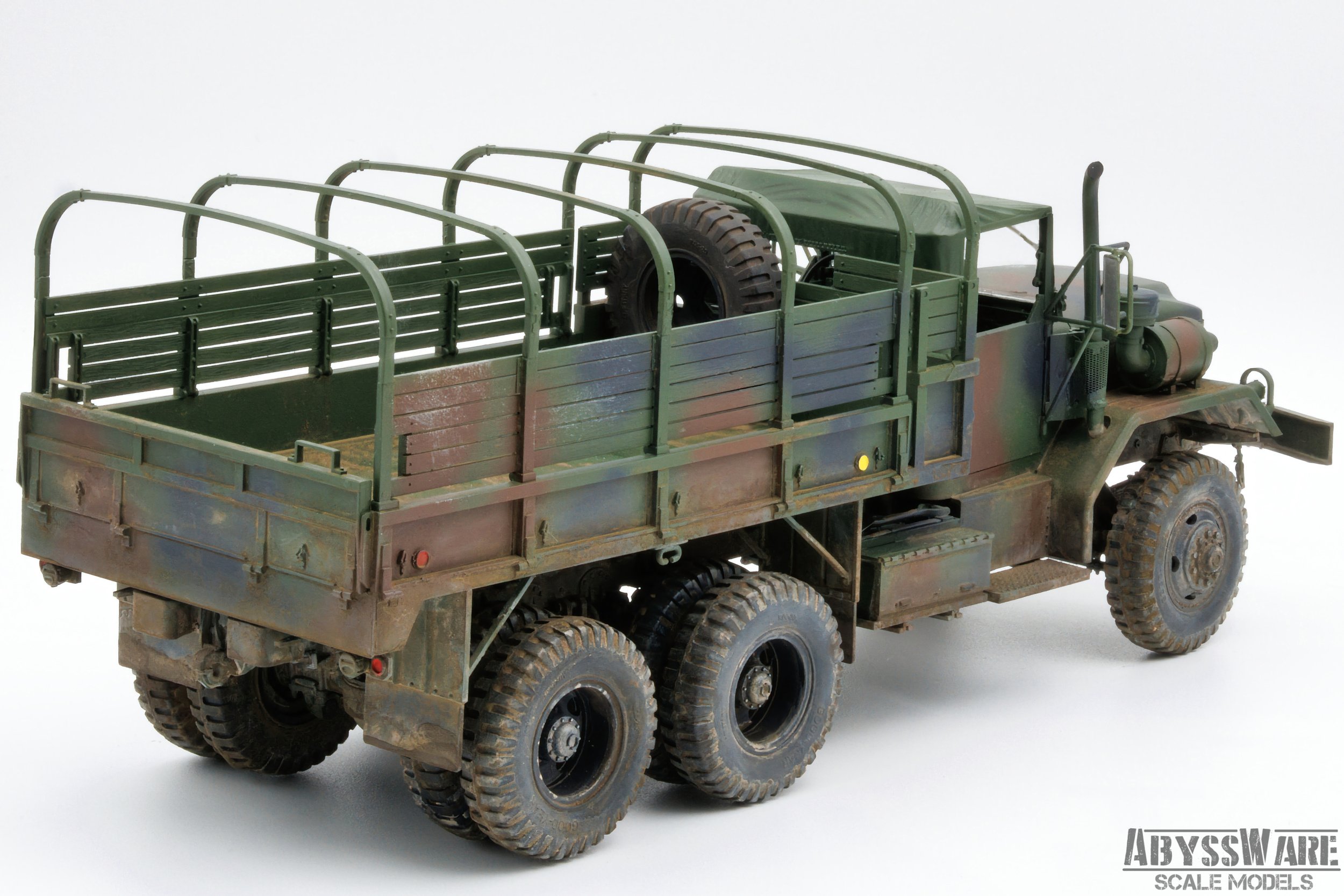 Scale model of a military transport truck with a camouflage paint job, large off-road tires, and an open cargo area with green railings, placed against a plain white background.