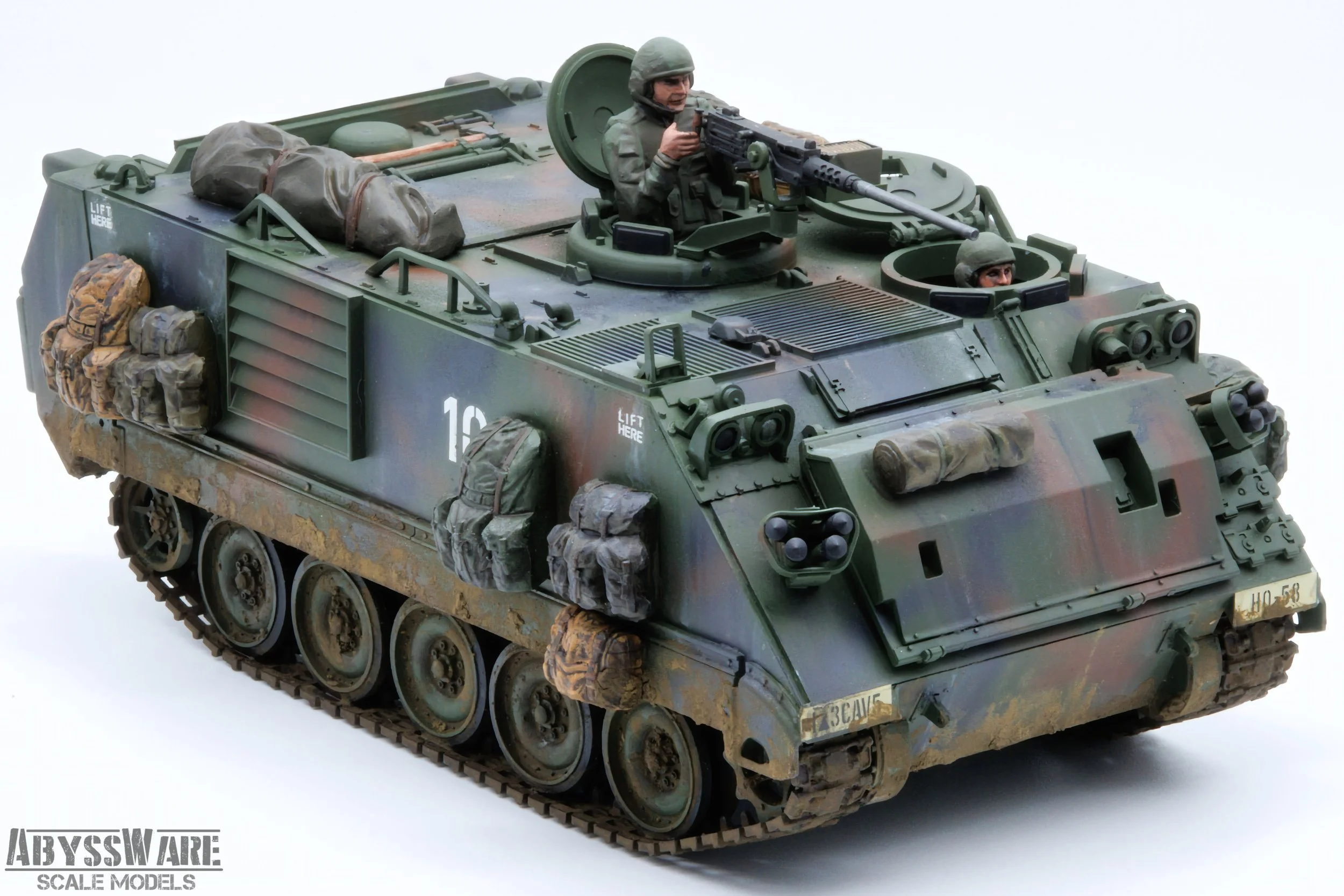 A detailed scale model of a military armored vehicle with camouflage paint, featuring two soldiers with helmets, one manning a machine gun and the other peeking out of a hatch, with backpacks and supplies attached to the vehicle.