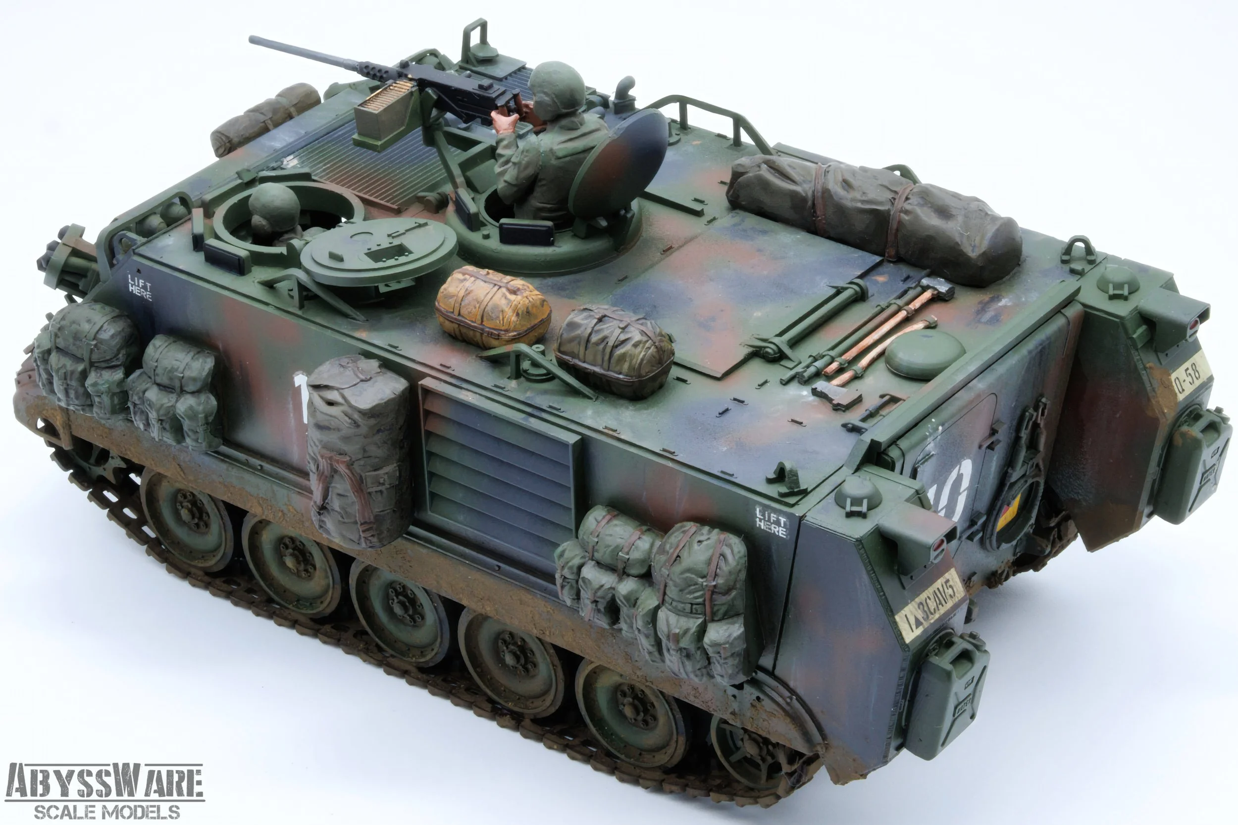 Scale model of a military armored vehicle with a soldier figurine aiming a machine gun, detailed with bags, equipment, and camouflage paint.