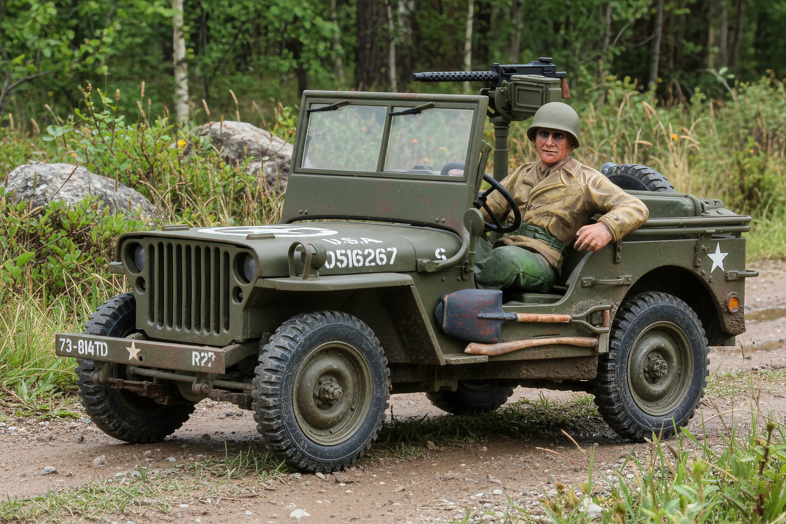 Model of a World War II military Jeep with a soldier figurine seated in the driver’s seat, outdoors on a dirt path surrounded by greenery.