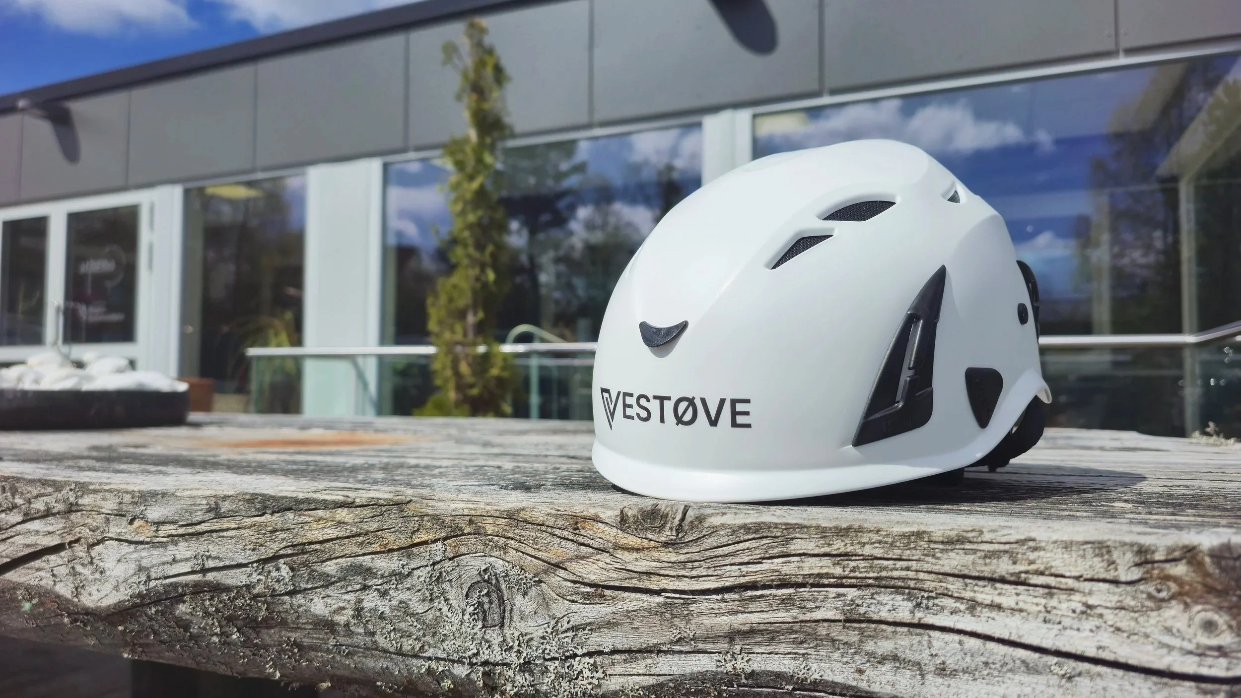 White cycling helmet with vent holes and black accents, placed on a weathered wooden surface outdoors, with a modern building and glass windows reflecting trees and sky in the background.
