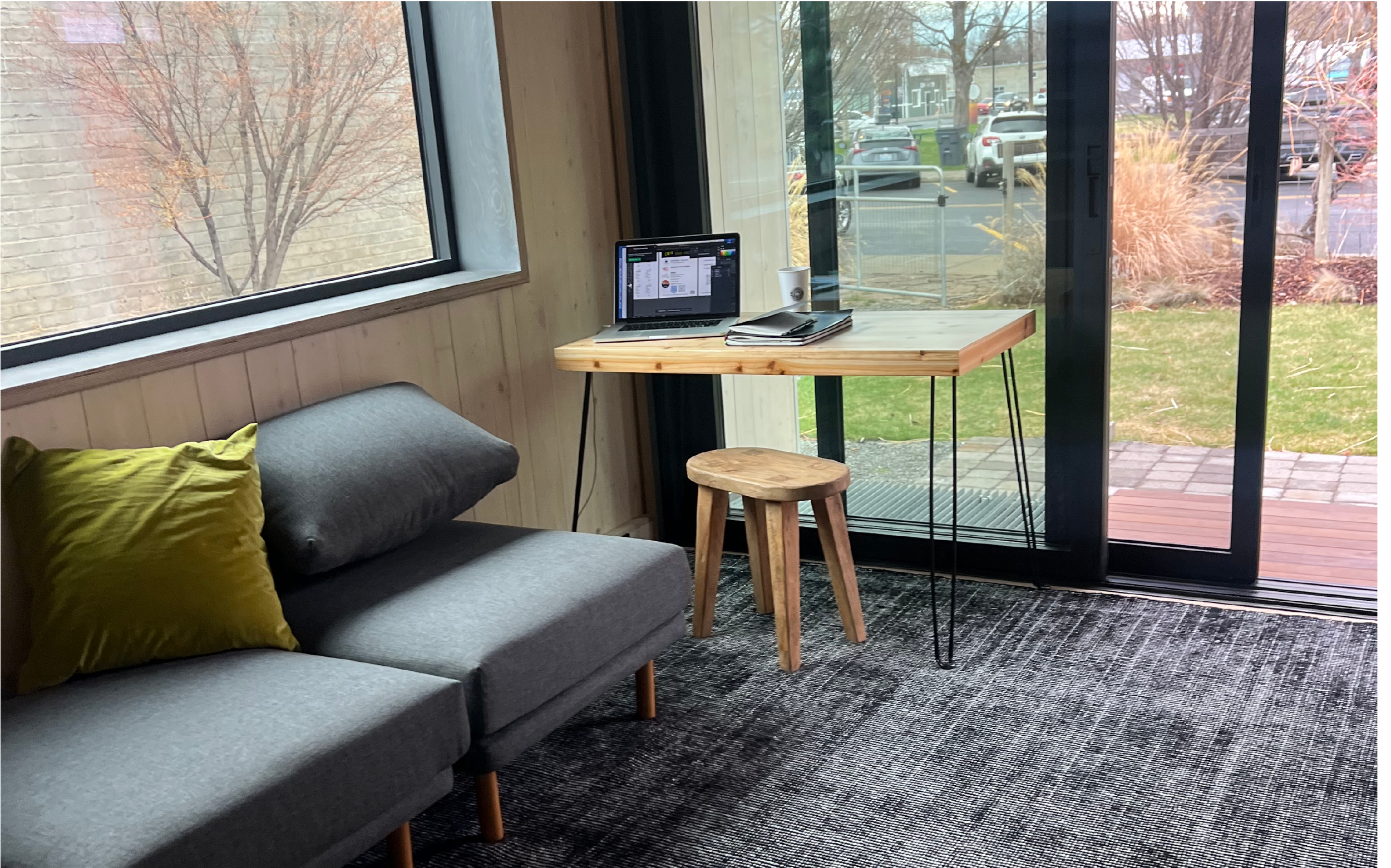 A cozy workspace with a grey couch accented by a yellow pillow, a wooden table holding a laptop, a coffee cup, and a book, near large glass windows and sliding glass doors outside showing a green lawn, trees, and parked cars.