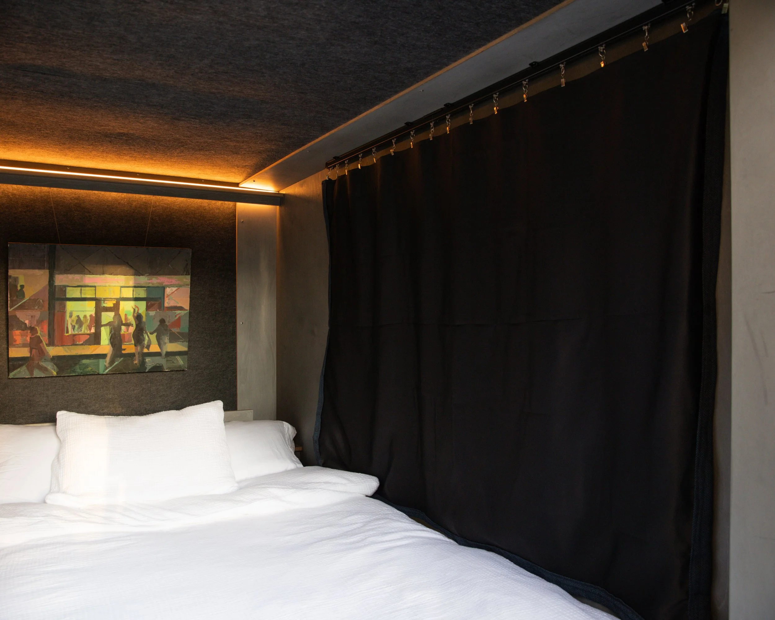 A cozy bedroom with white bedding, a dark wall, a dark privacy curtain, a colorful wall art, and soft lighting.