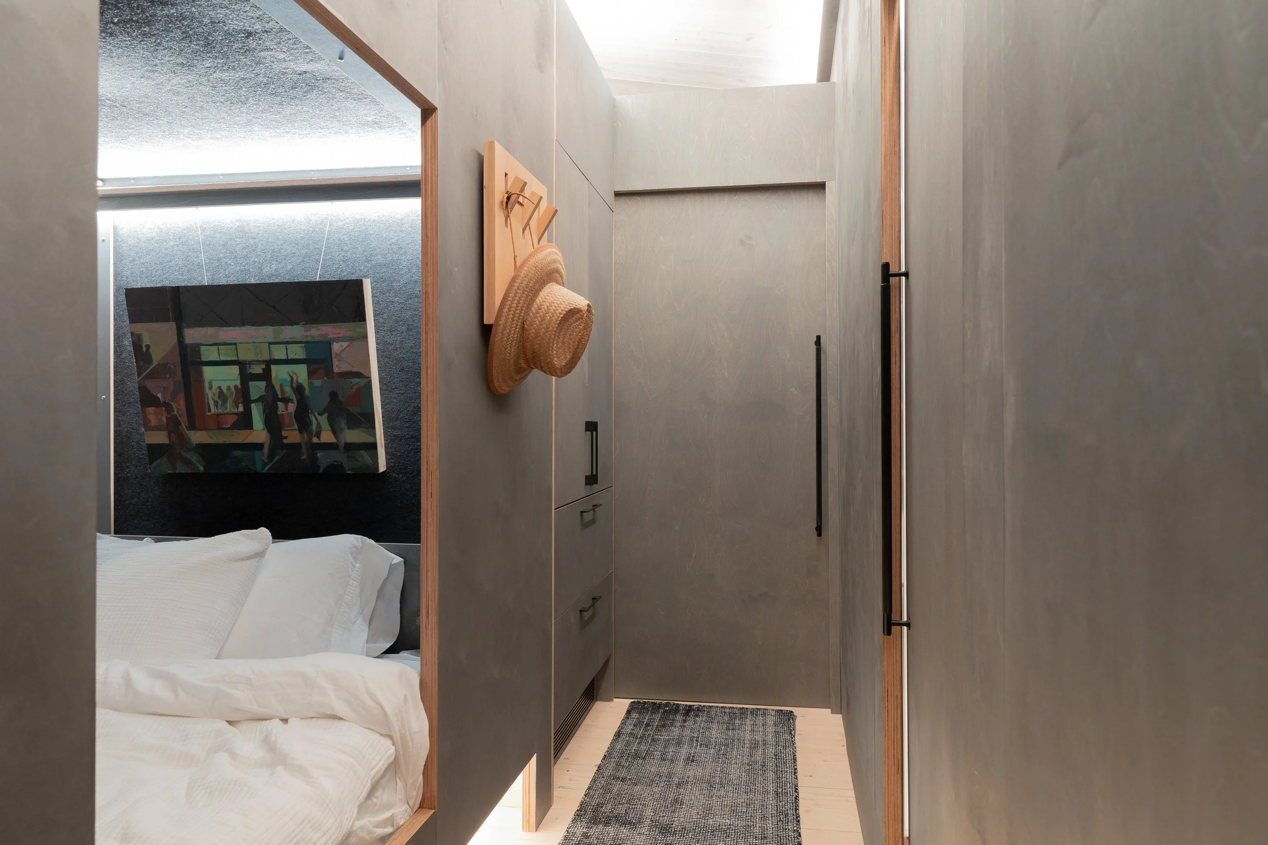 Interior view of a small bedroom with gray walls, a bed with white linens, a painting on the wall, and a hallway with a gray rug, cabinets, and hats hanging on hooks.