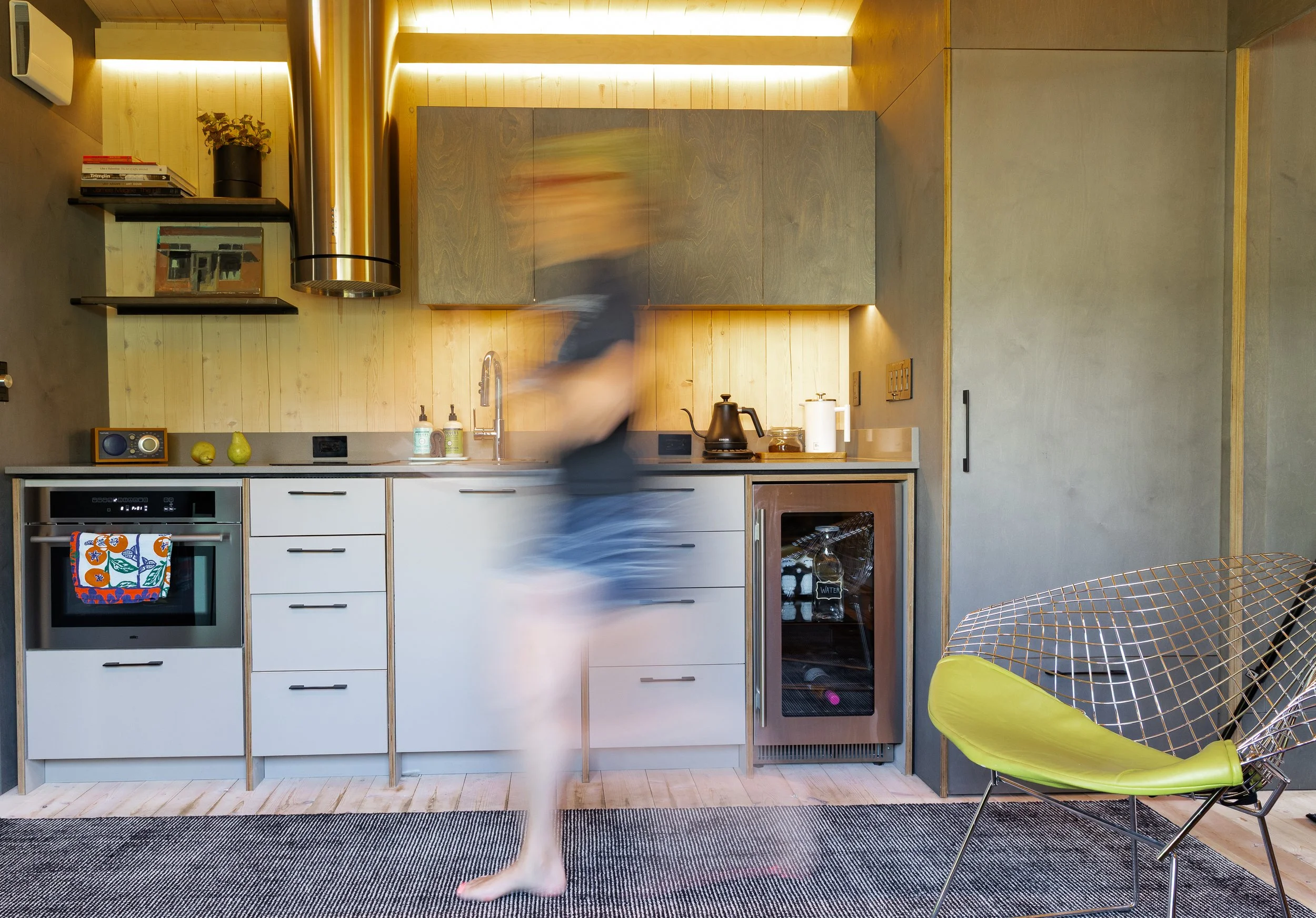Blurred person walking in a modern kitchen with white cabinets, a small wine fridge, a chair with a yellow cushion, and wooden accents.
