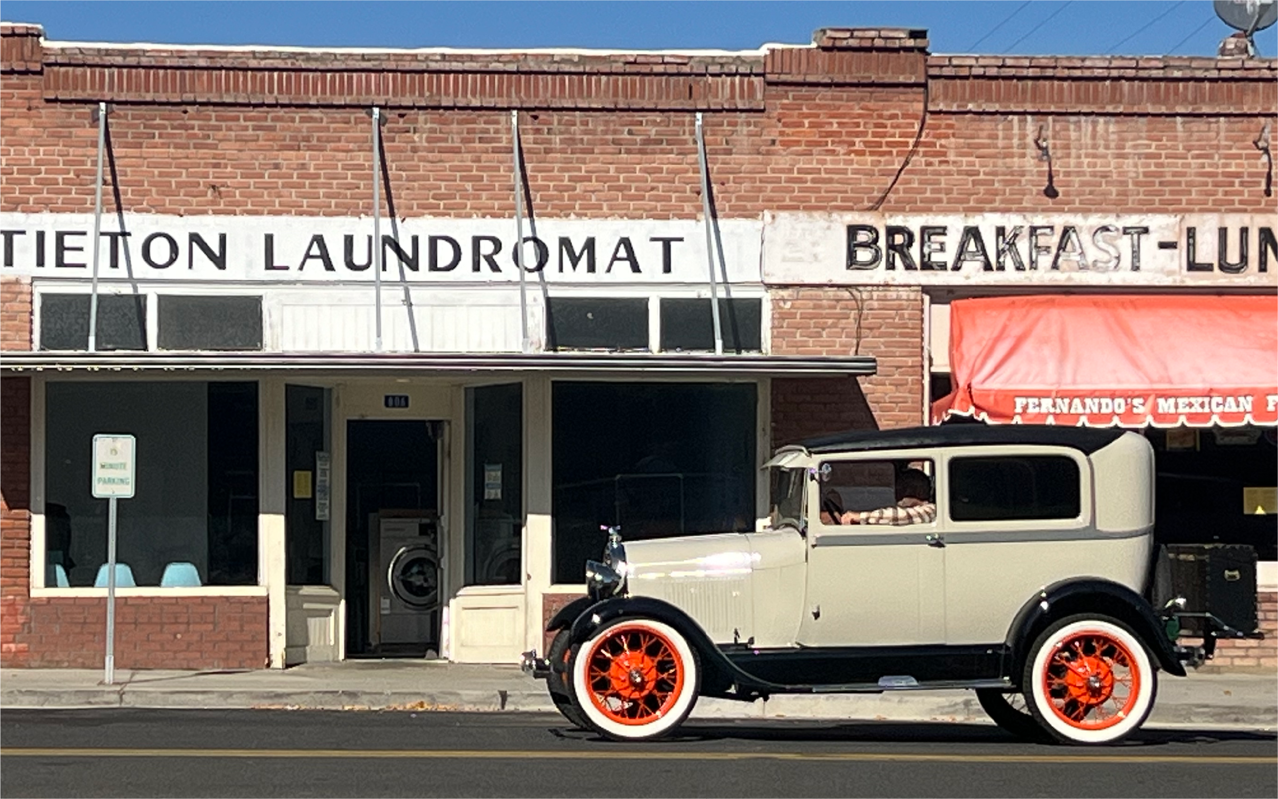 A vintage car with cream body, black accents, and orange wheels parked in front of a brick building with signs reading 'Laundromat' and 'Breakfast Lunch'.