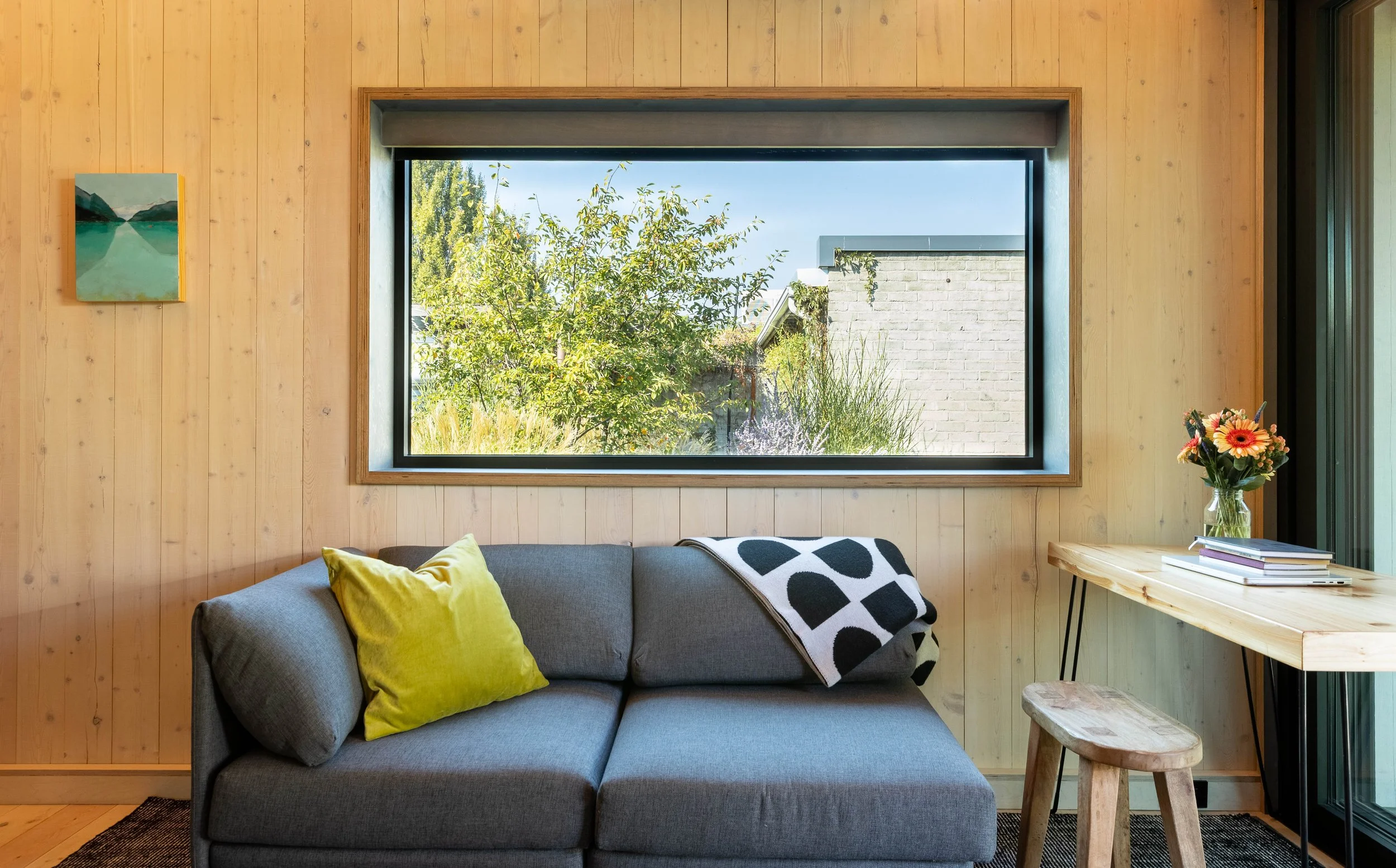 Interior of a cozy living room with a gray sofa, a yellow pillow, a black and white patterned blanket, a wooden table with flowers and books, and a large window showing greenery outside.