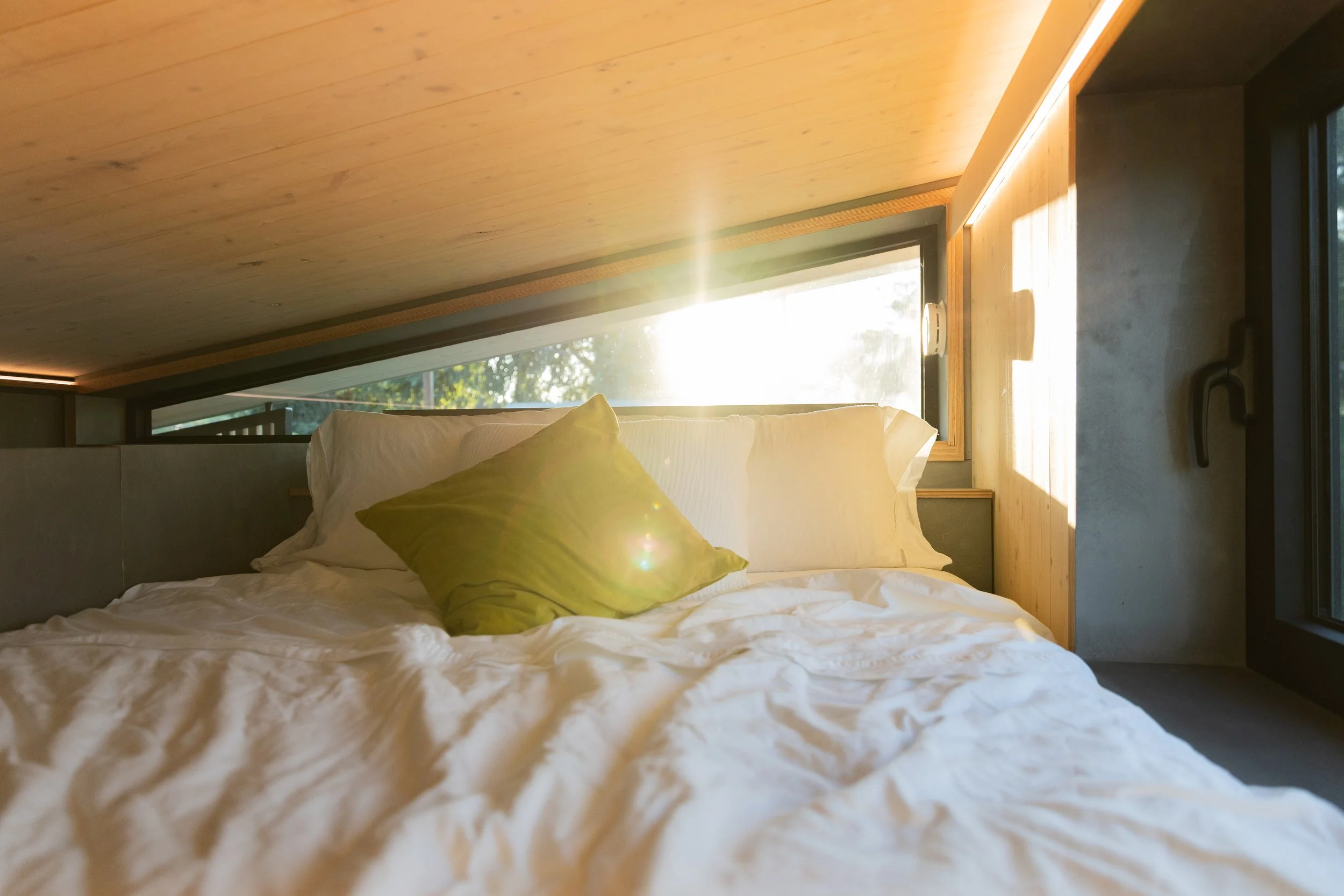 A bed with white bedding and a green pillow in a room with a large window letting in sunlight, wooden ceiling, and concrete wall.