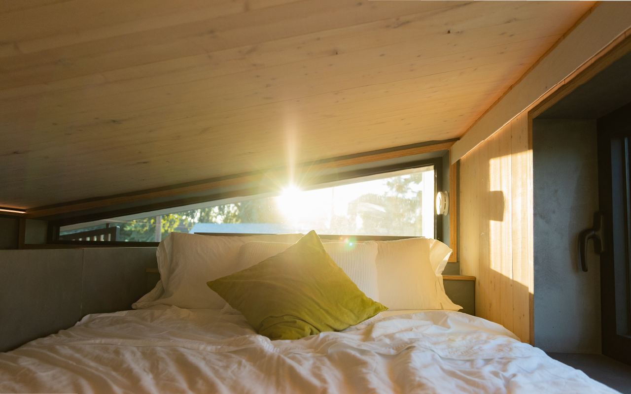 Sunlight shining through a window onto a bed with white bedding and a green pillow in a cozy room with wood-paneled ceiling and walls.