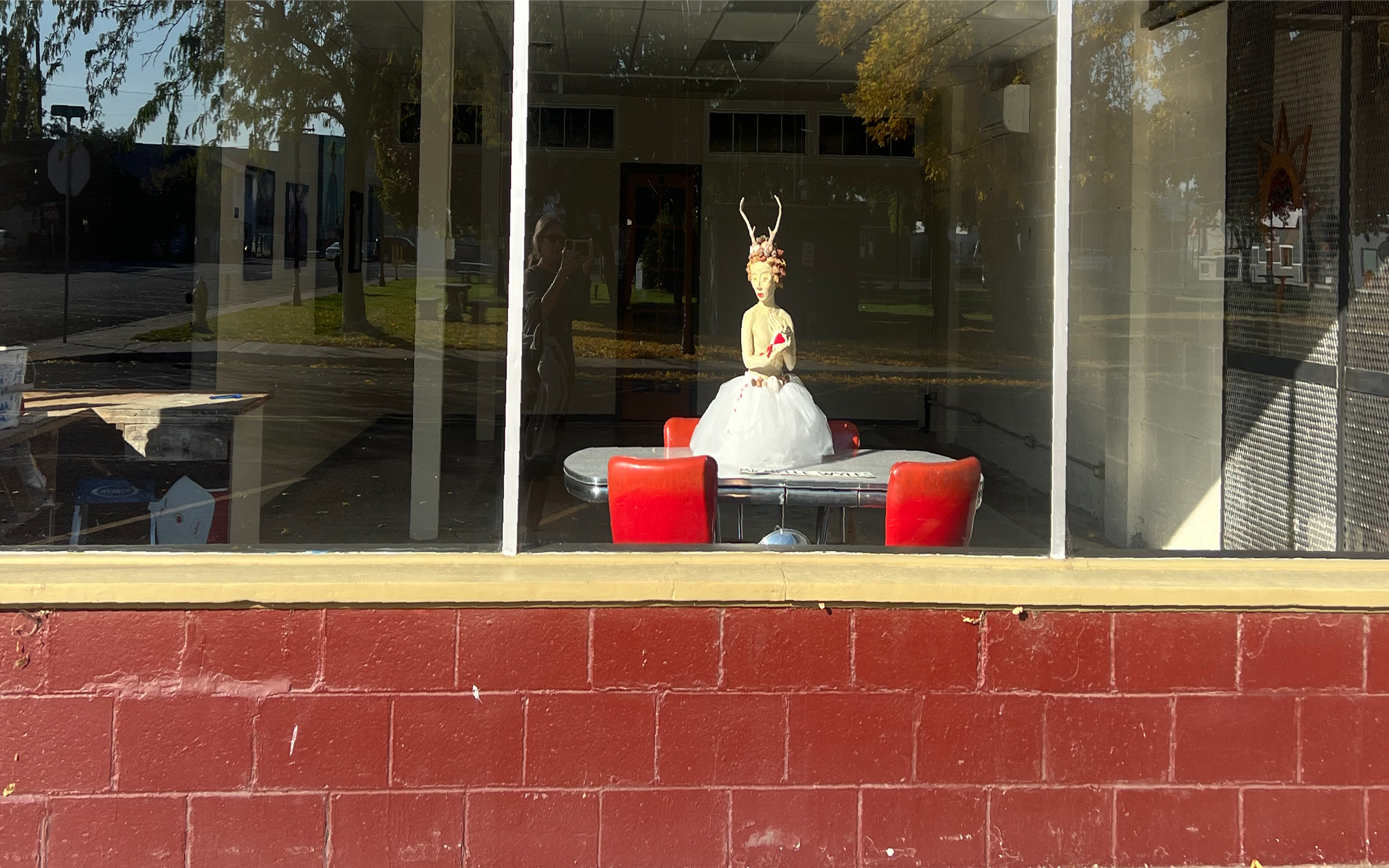 a display of a figurative sculpture with an intricate twisted horn headdress and a white dress, sitting on a round table with red chairs around it, is seen through a glass window with reflections of outside trees and buildings.