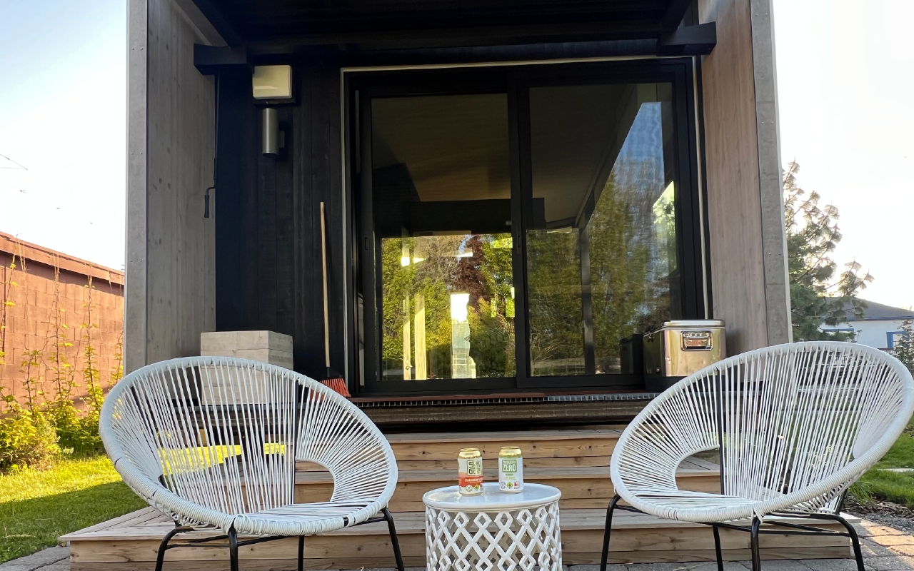 Two white circular outdoor chairs with a small white table between them, holding two cans of beverages, in front of a modern glass door leading into a house.