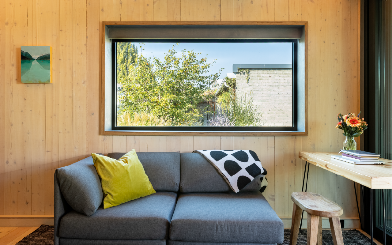 A cozy living room with wood-paneled walls, a large window showing greenery outside, a gray sofa with a green pillow and a black and white throw blanket, a small wooden table with a vase of flowers and books, and a small wooden stool.