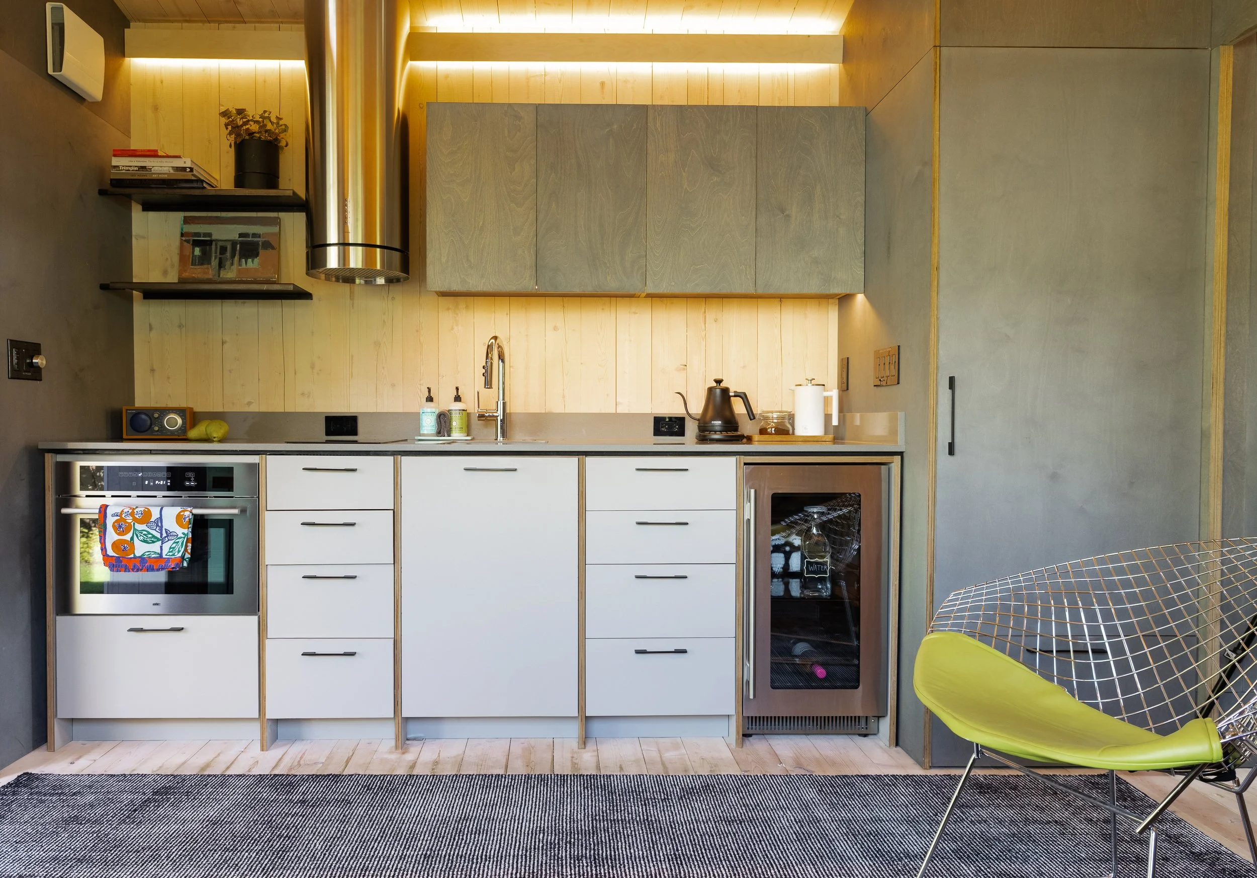 Modern kitchen with white cabinets, wooden backsplash, stainless steel appliances, black kettle, small wine refrigerator, and a wireframe chair with lime green cushion in front of a textured gray wall.