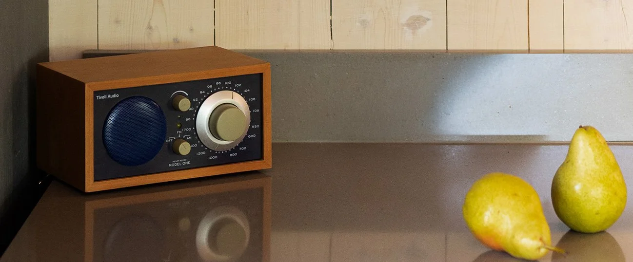 A vintage brown wooden speaker with control knobs and a large tuning dial on a reflective brown surface, next to two yellow pears on a gray countertop against a wooden wall background.