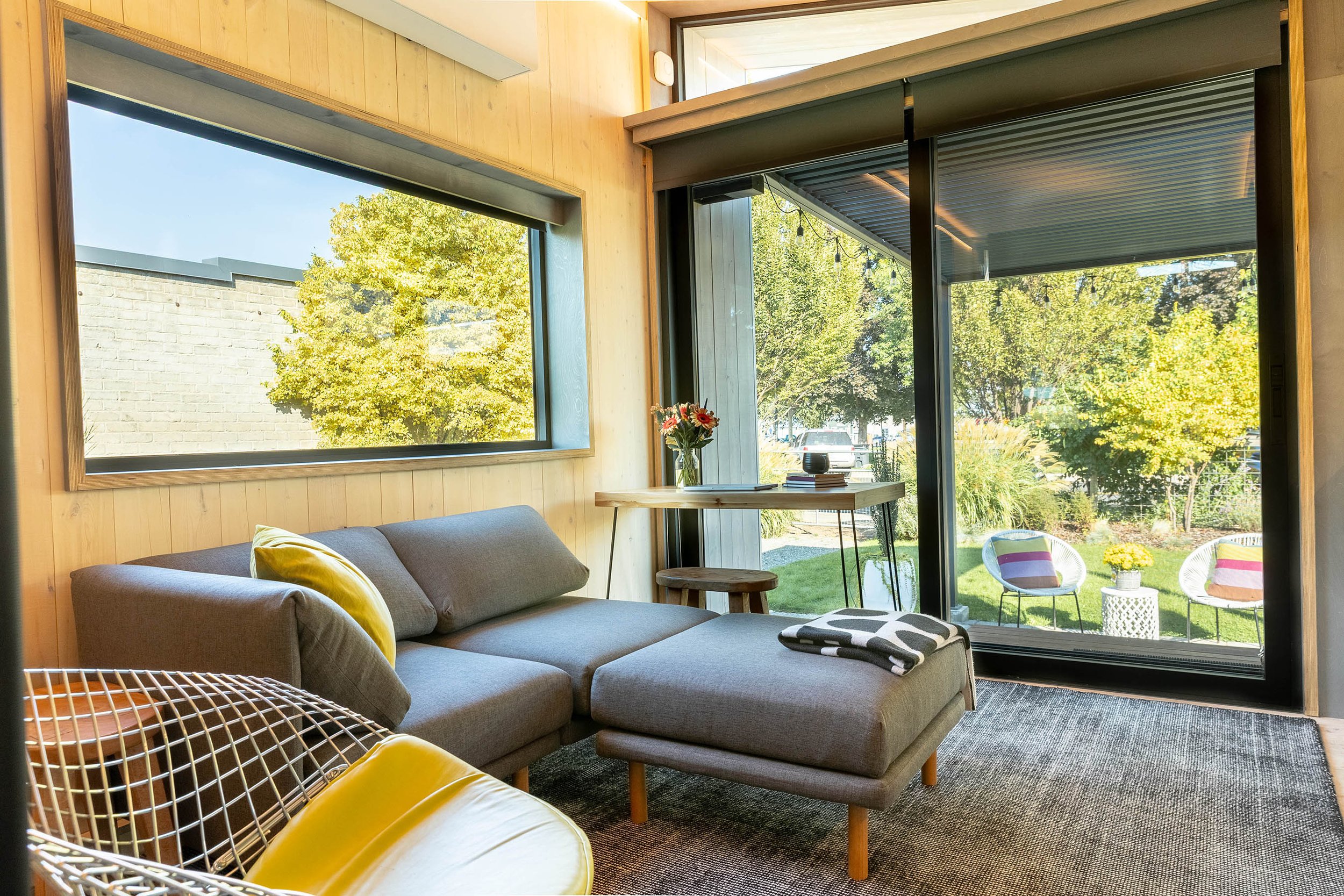 Living room with gray sectional sofa, window and sliding door leading to outdoor patio with two chairs, small table with yellow flower pot, and view of garden.