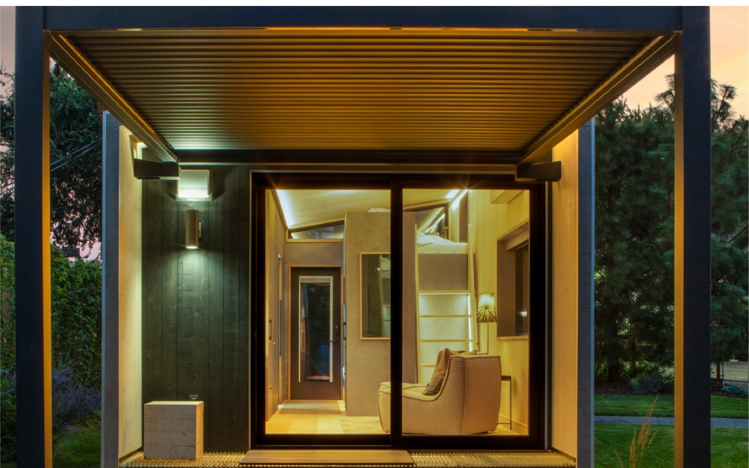 Interior view of a modern small house with large glass doors, warm lighting, a beige armchair, and a loft bed visible through the glass.