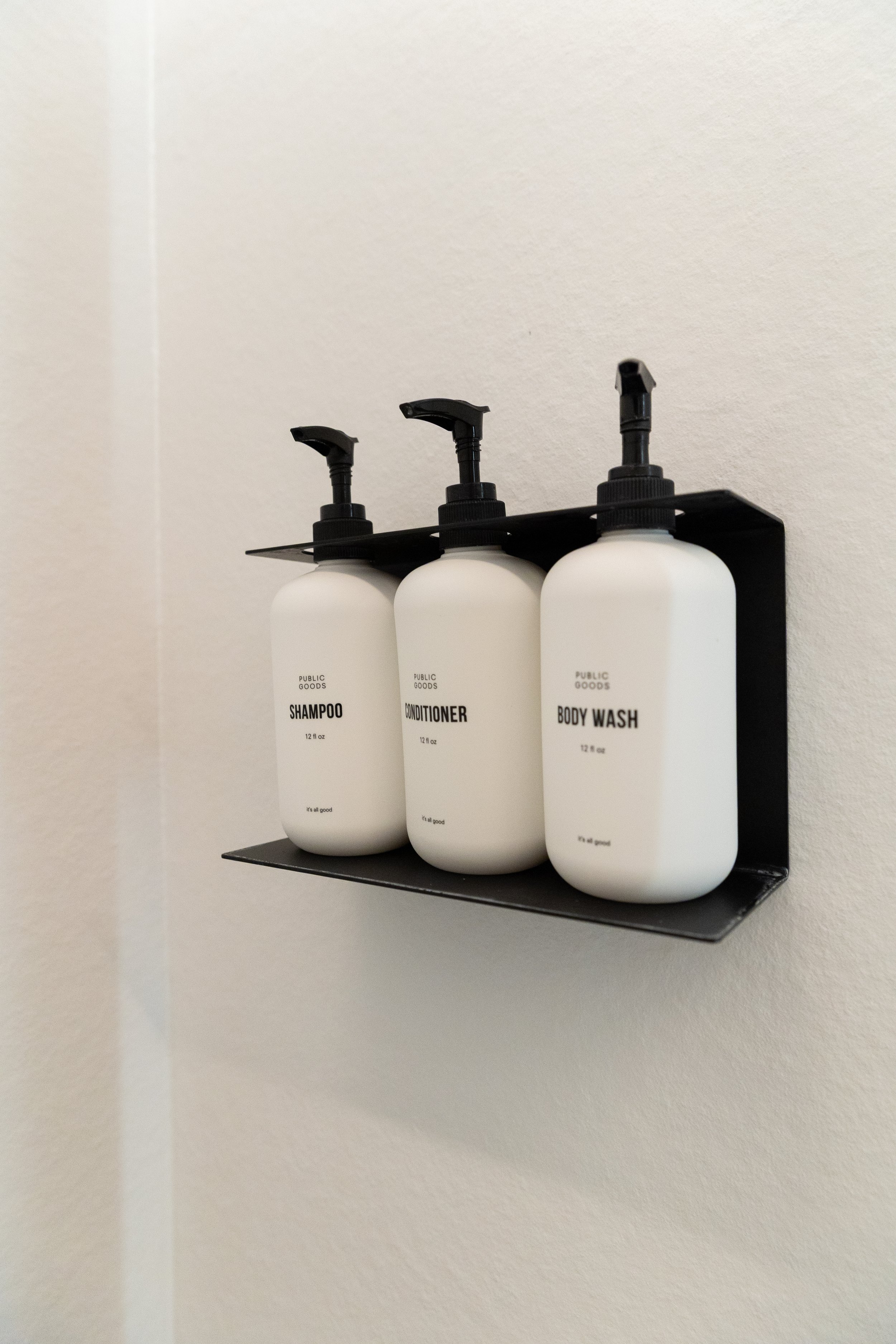 Black shelf mounted on the wall holding three white bottles labeled shampoo, conditioner, and body wash with black pumps.