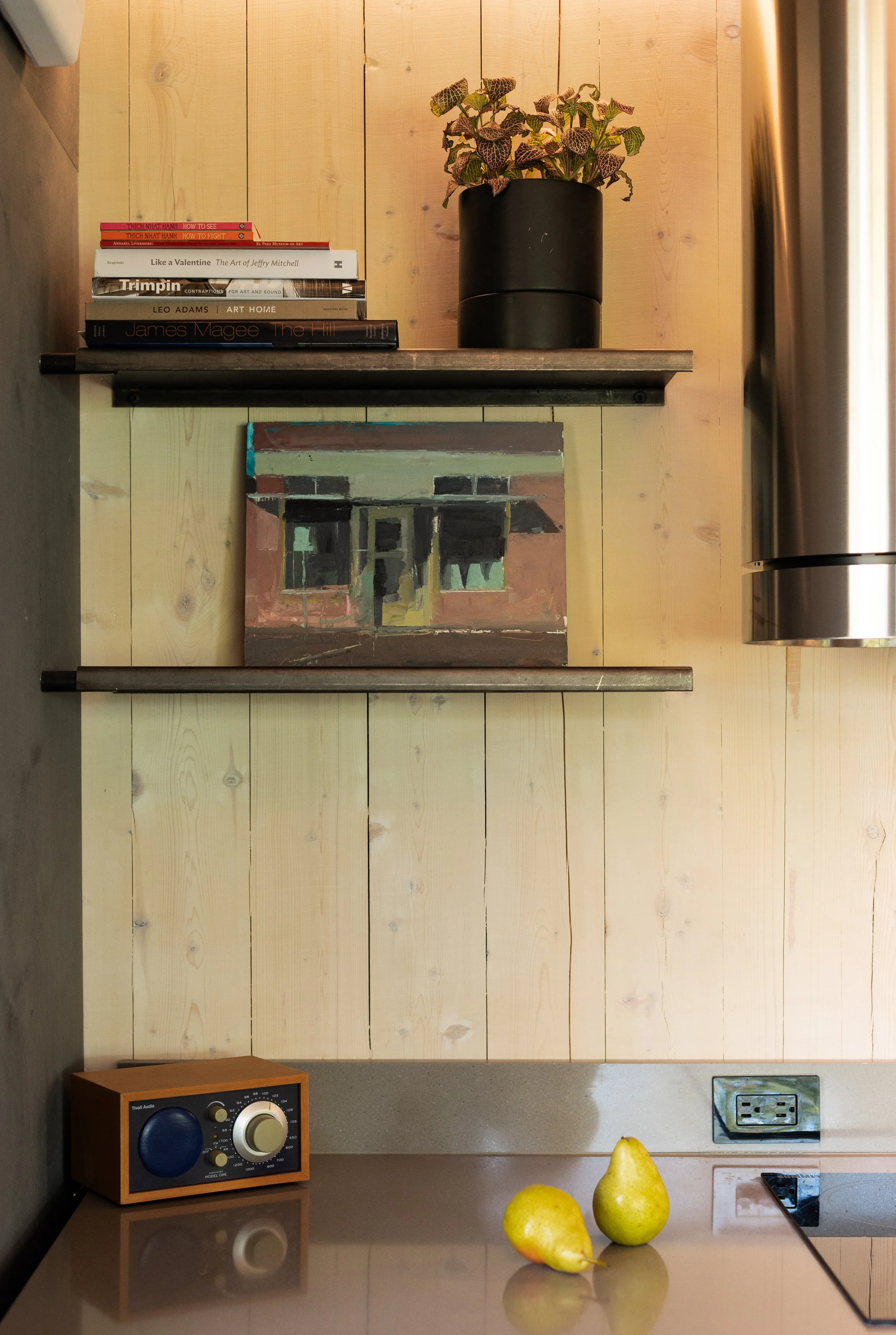 A kitchen corner with light wood paneled wall, a black planter with patterned leaves on a wood shelf, a stack of books including art and design topics, a small abstract painting, yellow pears on the counter, a vintage radio, a power outlet, and a sta