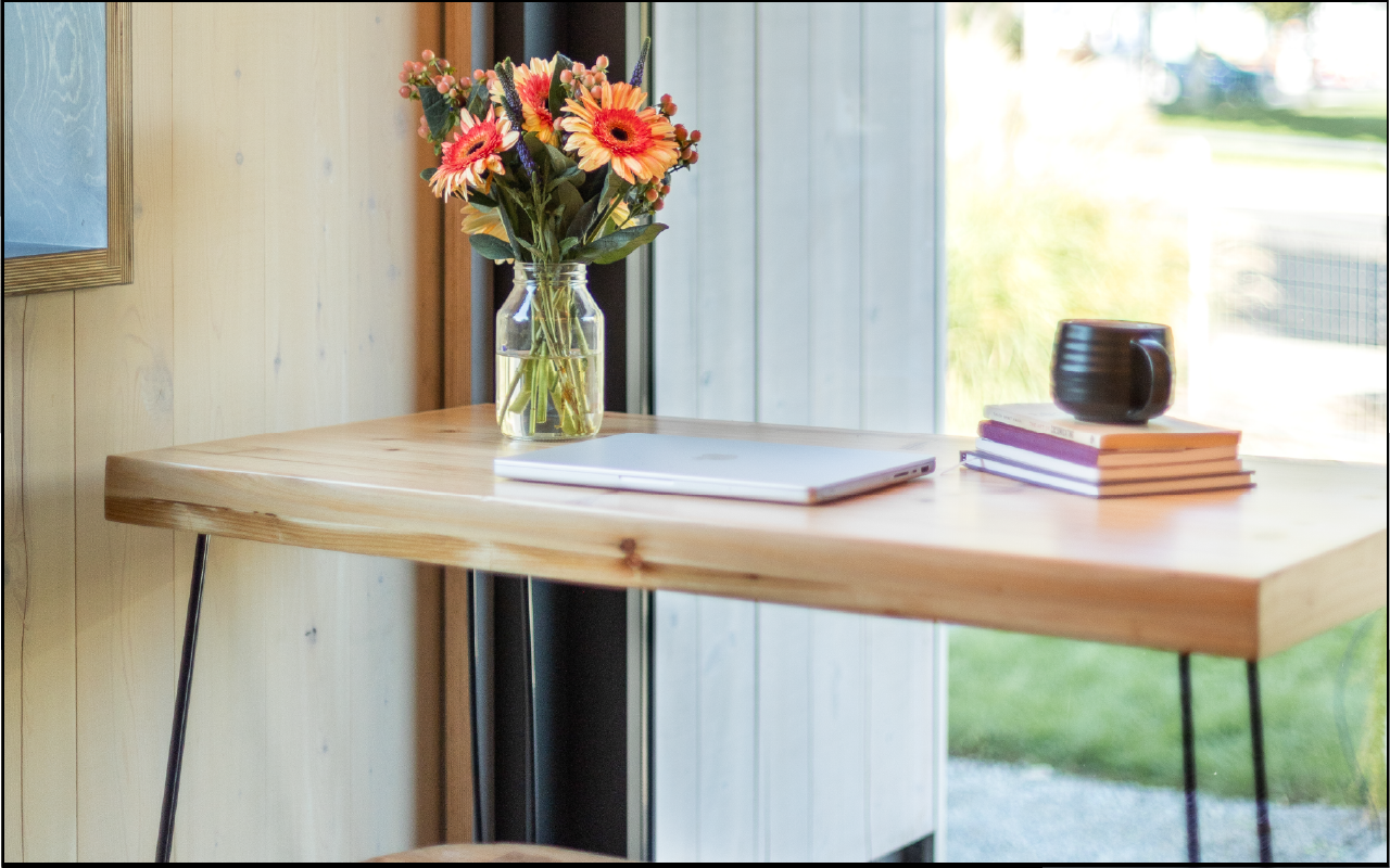 A wooden desk near a window with a vase of flowers, a closed laptop, a stack of books, and a black mug.