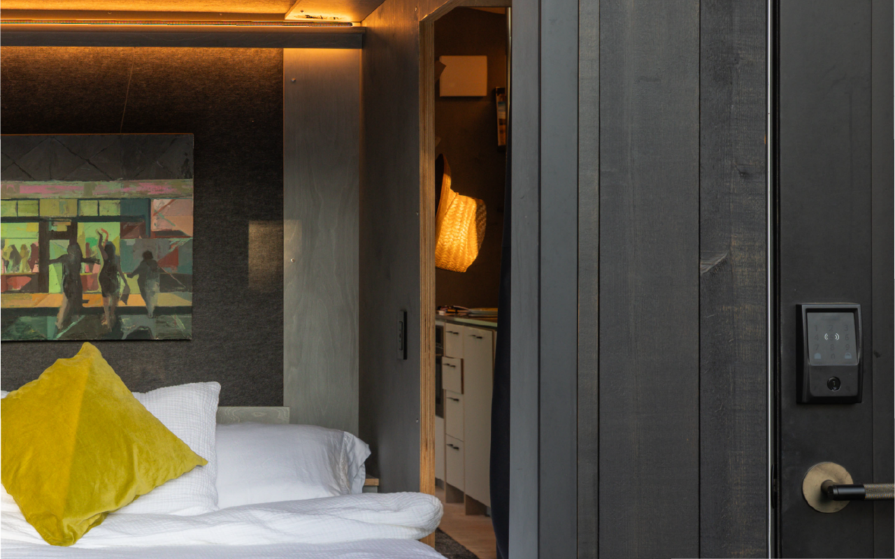 Part of a bedroom with a bed, yellow pillow, and artwork on the wall, with a sliding door leading into a kitchenette area.