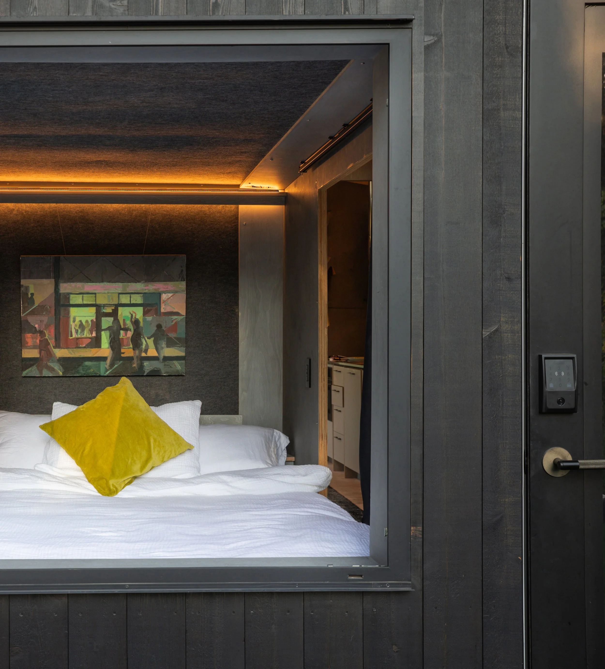 A view of a modern bedroom seen through an open window or hatch. The bed has white bedding and a yellow pillow. Artwork hangs above the bed, and a hallway or kitchen is visible in the background.