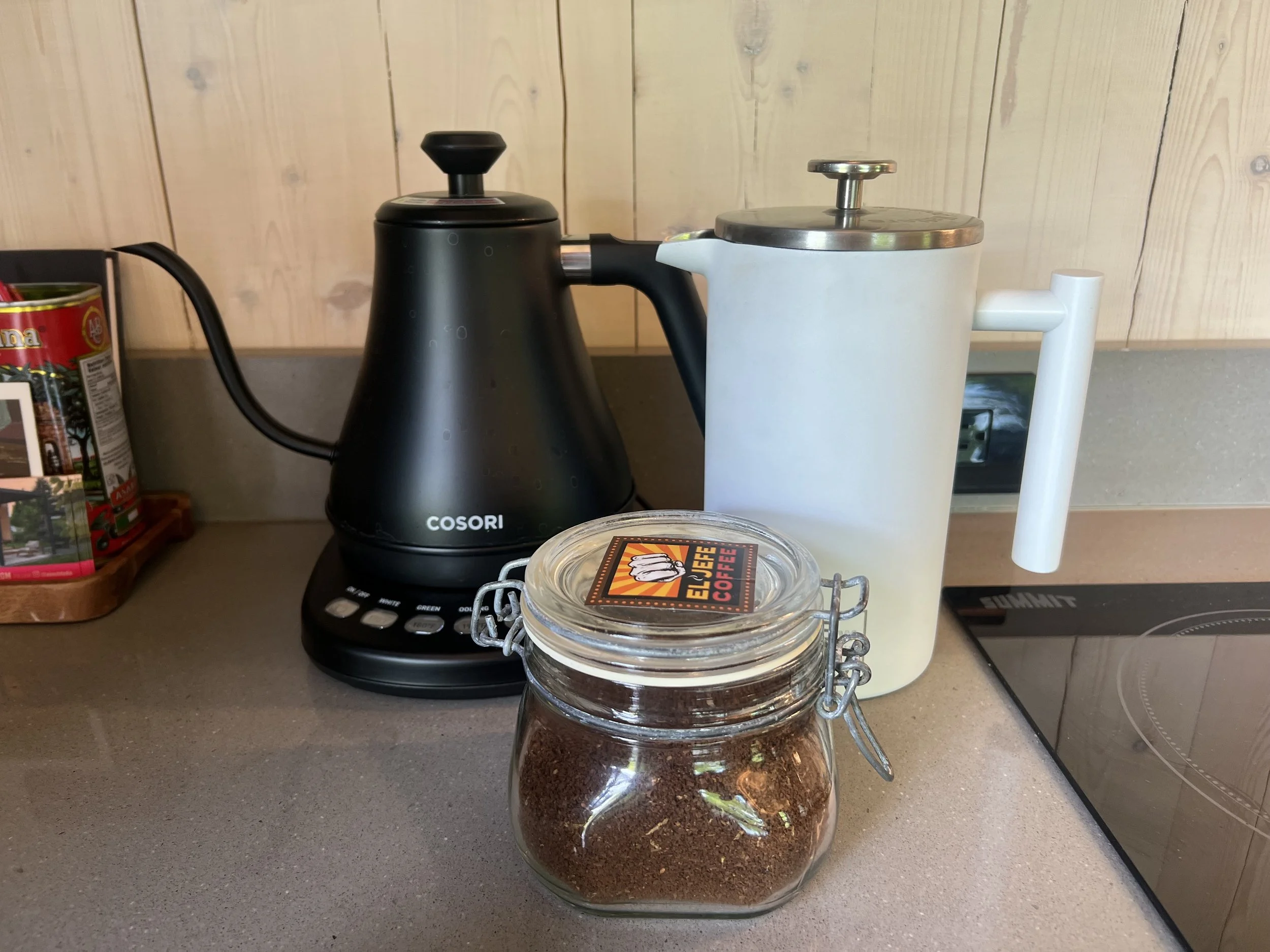 A black electric kettle, a white French press, and a glass jar of ground coffee on a kitchen countertop with a wood-paneled wall in the background.