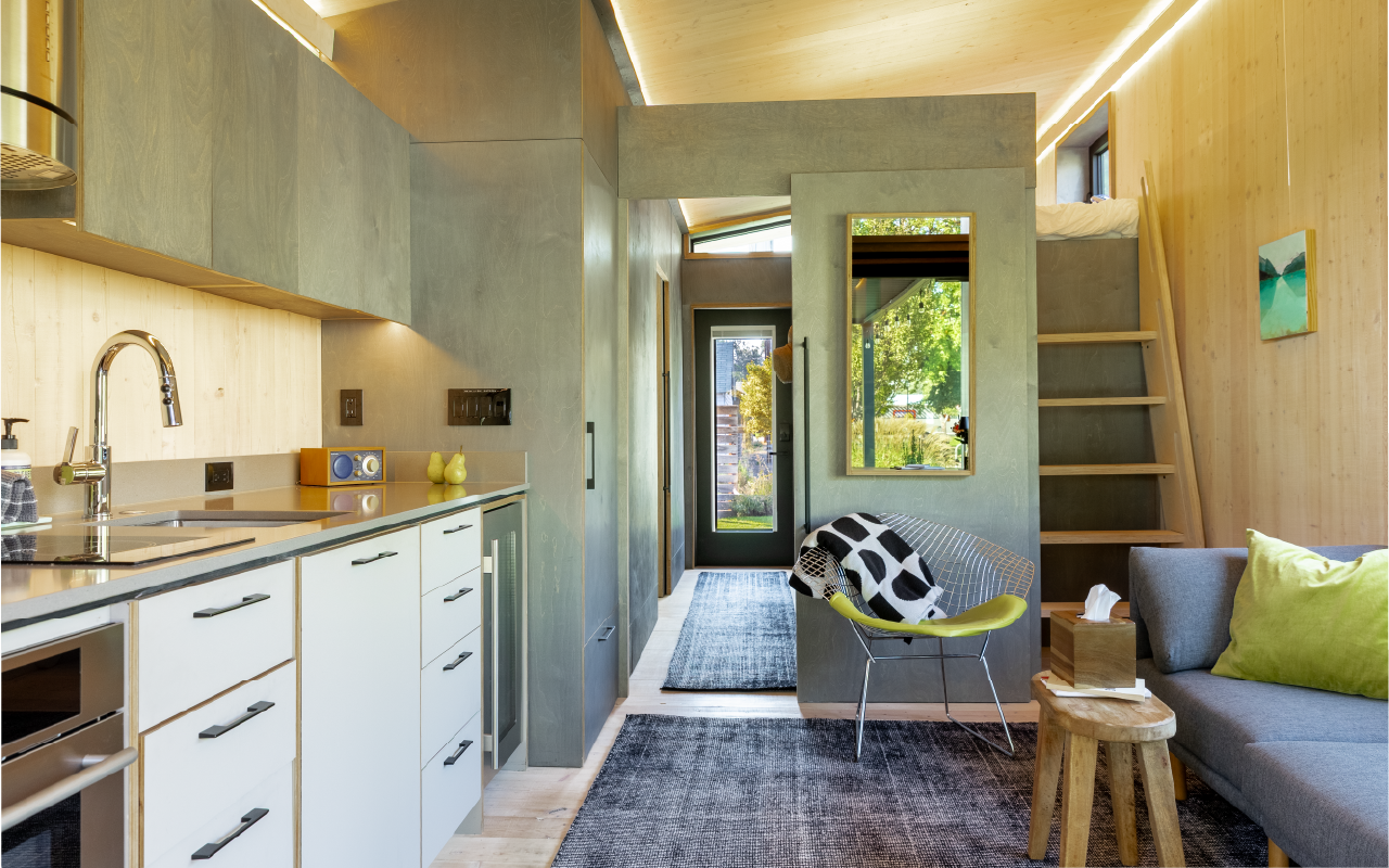 Interior of a modern tiny home with a compact kitchen on the left, a seating area with a gray sofa and a lime green pillow on the right, and a small staircase leading to a loft bed at the back. There is a door with a window and a mirror on a partitio